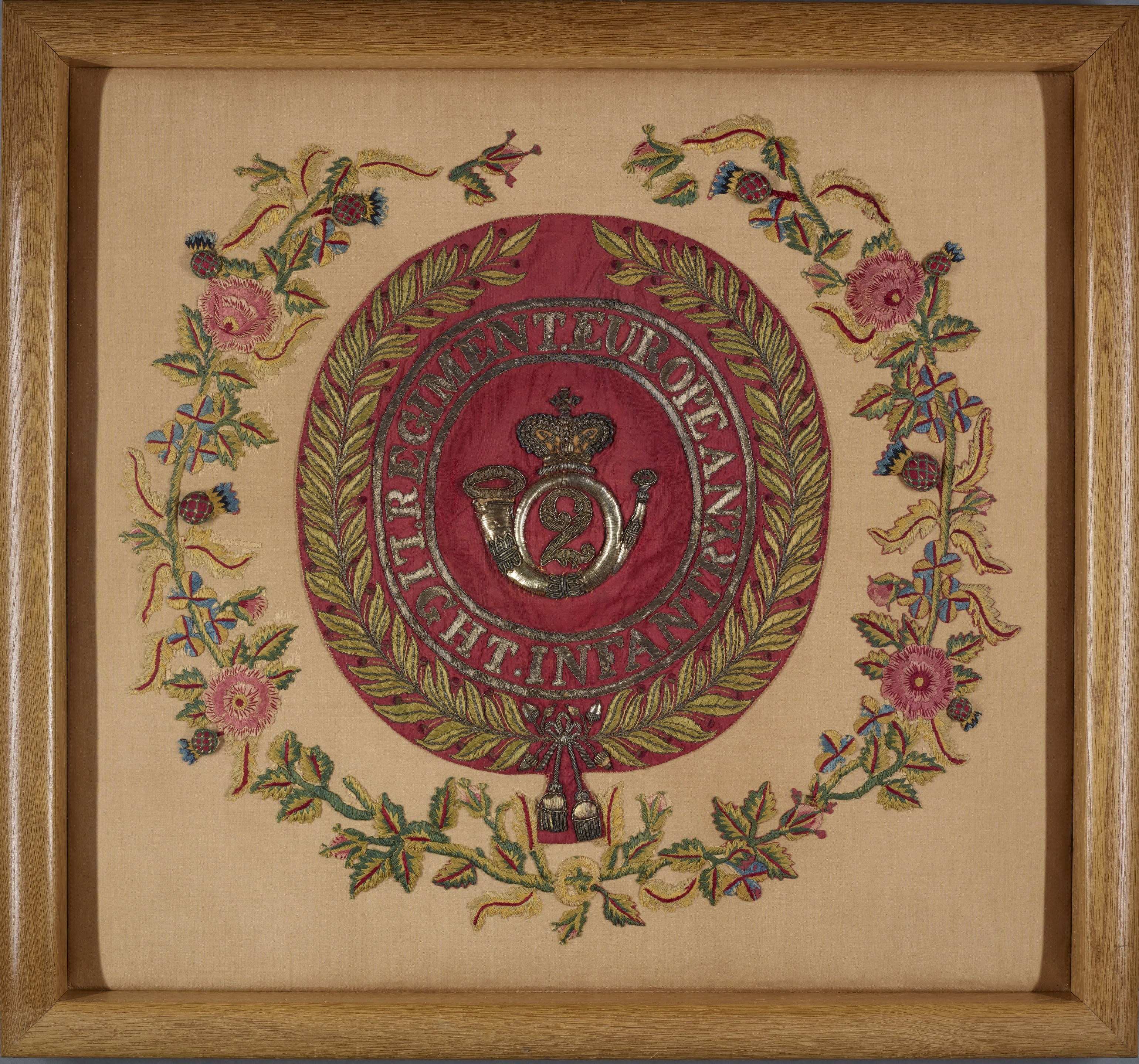 Photo of the Colour of the 2nd Bombay Light Infantry, in a wooden frame. The central design is red, with bugle logo in silver. The surrounding wreath is green leaves with pink flowers.