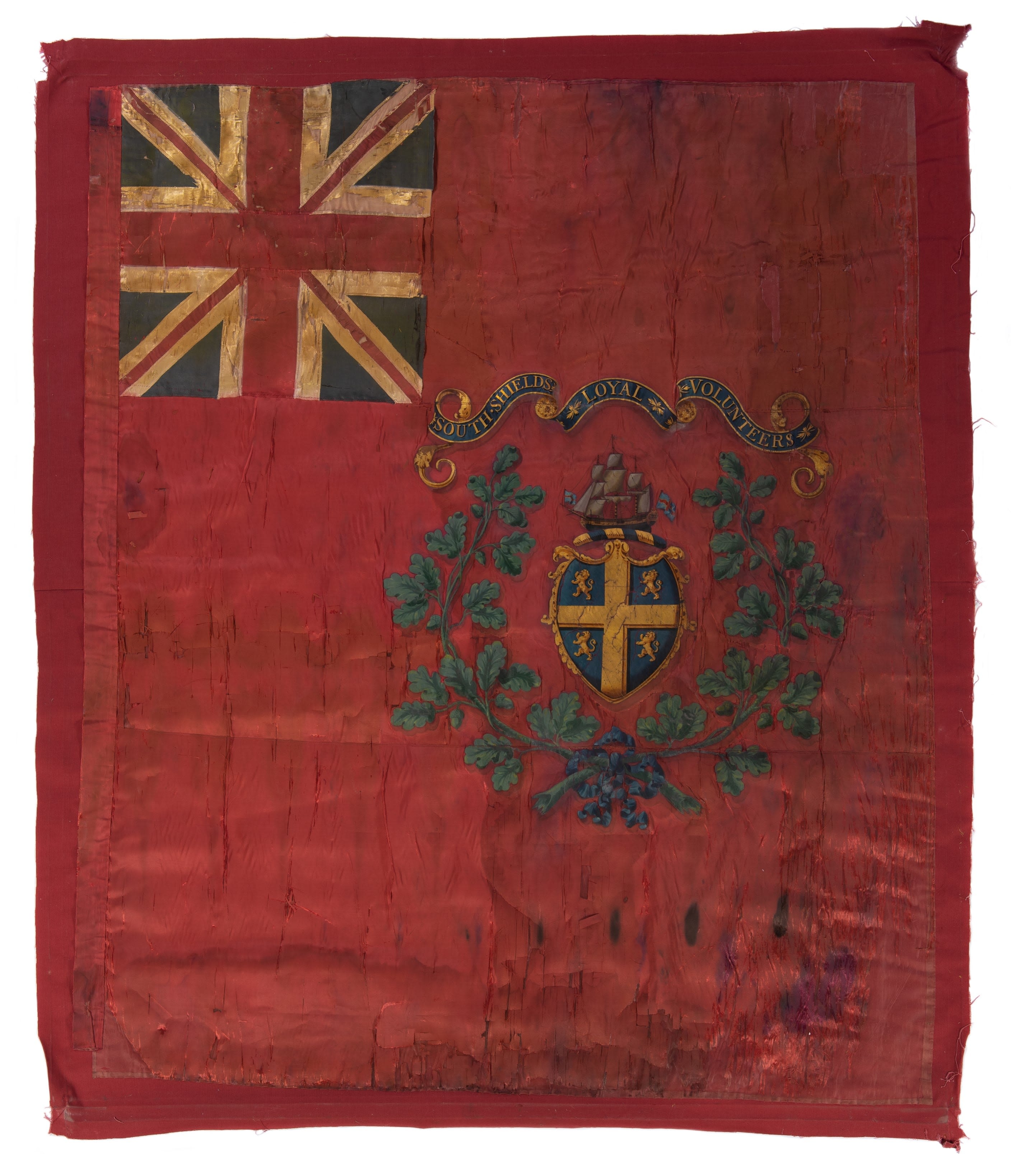This Colour has a red silk, background, with dark coloured streaks caused by age. The top left corner of the Colour has a Union Jack and there is a crest in the centre with the name of the unit, a green wreath, a large ship and a golden cross on a blue background.