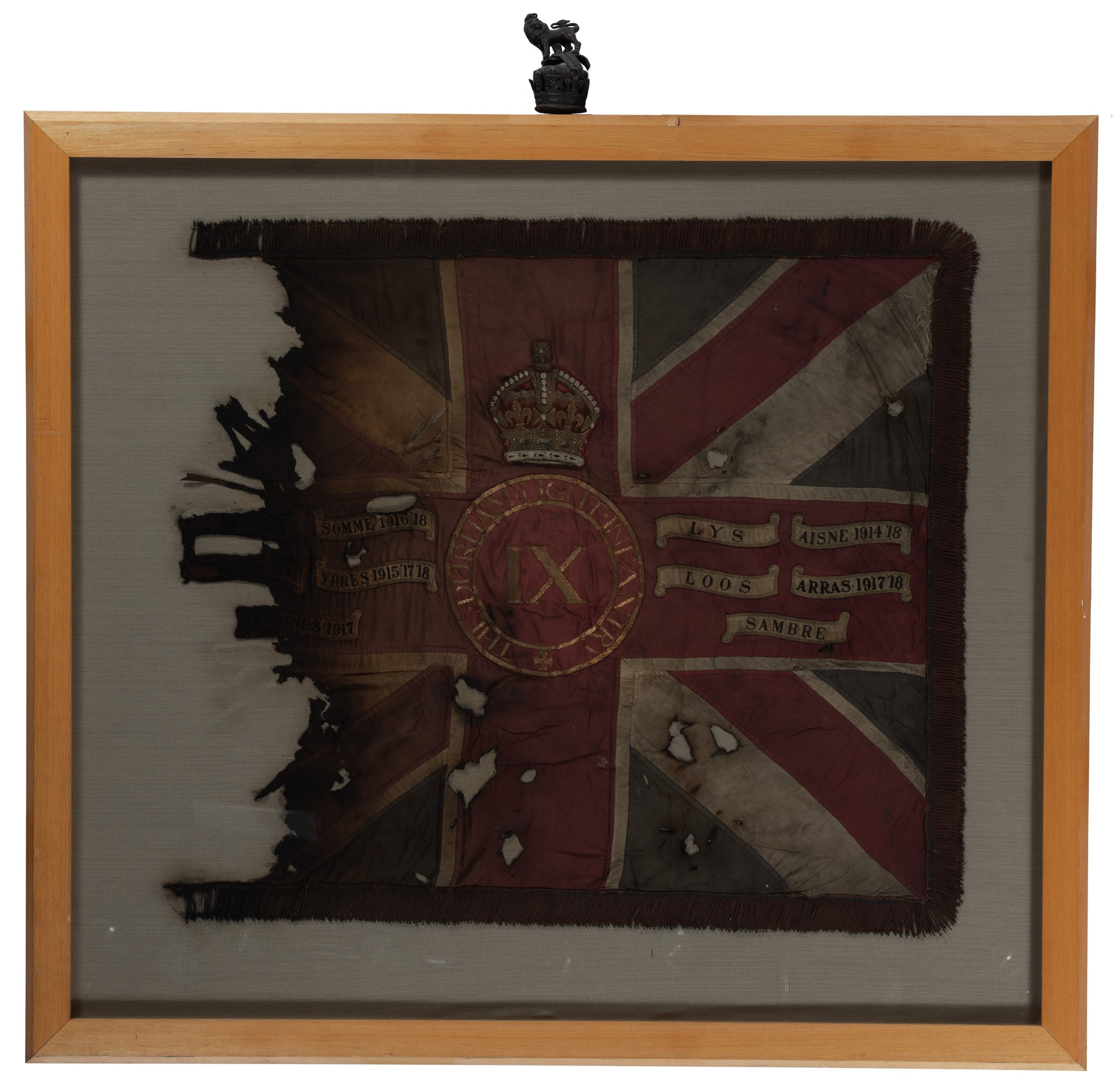 Photo of the King's Colours of 9 DLI in a wooden frame. The colour has significant fire damage, especially on the left hand side, which is almost completely disintegrated. Despite the damage, the Union flag and central crest of 9 DLI is still visible, along with some of the battle honours.
