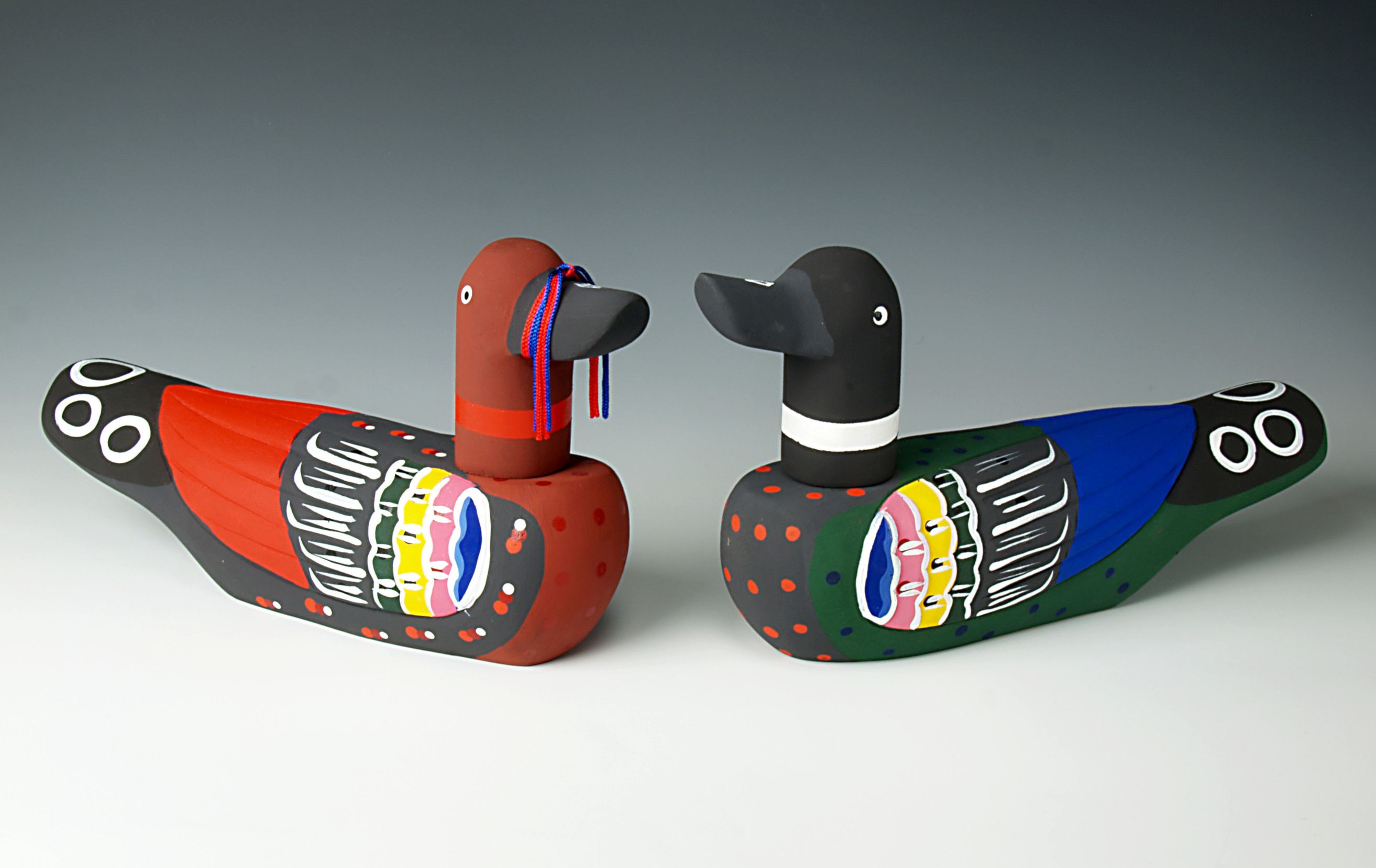 Two wooden wedding ducks sat face to face. One duck is red and black with a ribbon around its beak, the other is black and blue