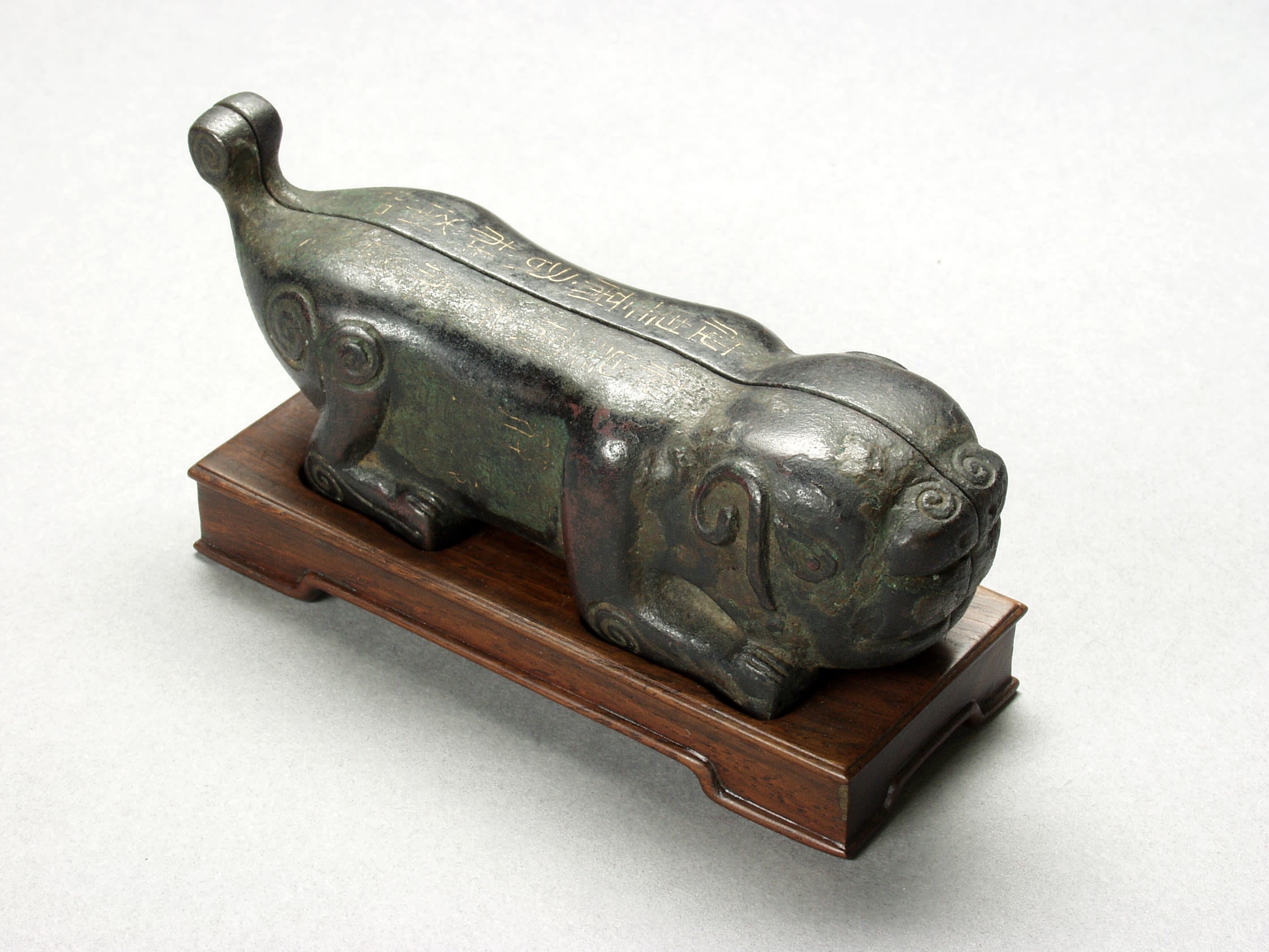 Sculpture on a wooden in the shape of a crouched tiger. The sculpture is split down the centre where it divides into two pieces. The sculpture is decorated with faint Chinese script and swirling patterns.