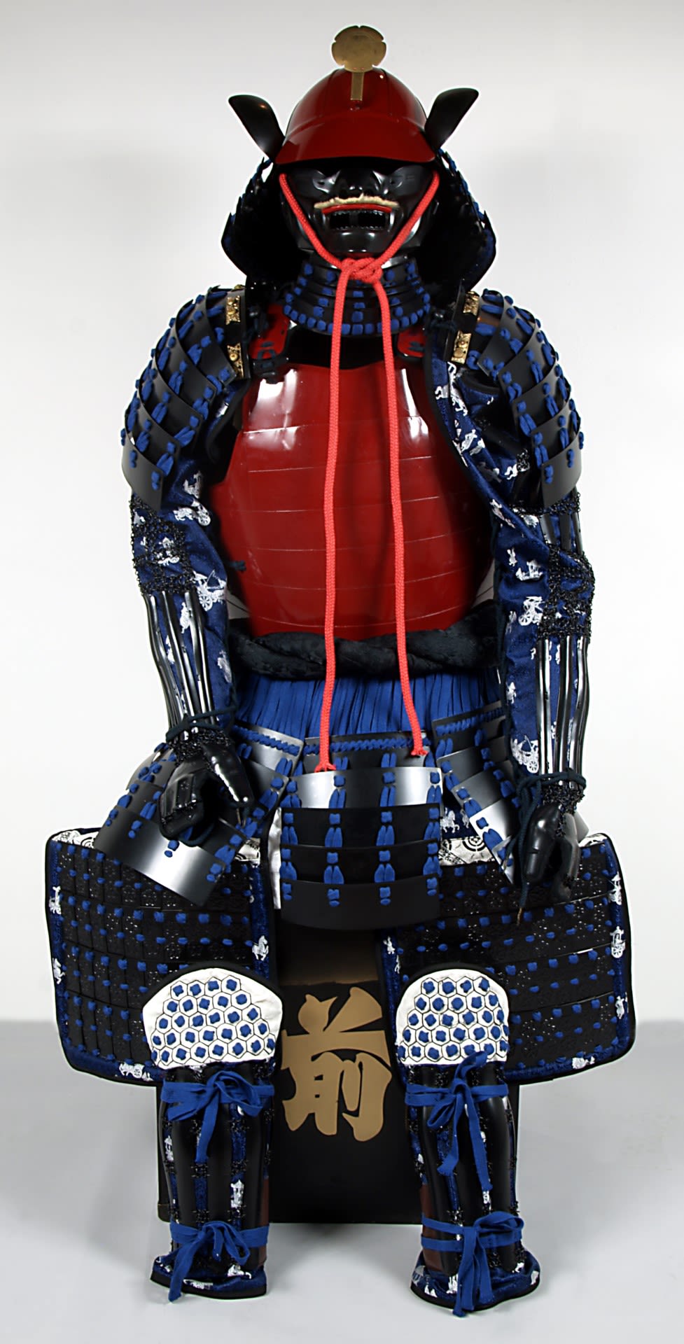 Blue, red and black samurai armour, combining sections of metal plate and scales which are laced together. The components of the armour are chest armour, helmet, facial armour, armoured sleeves, thigh armour and shin armour.