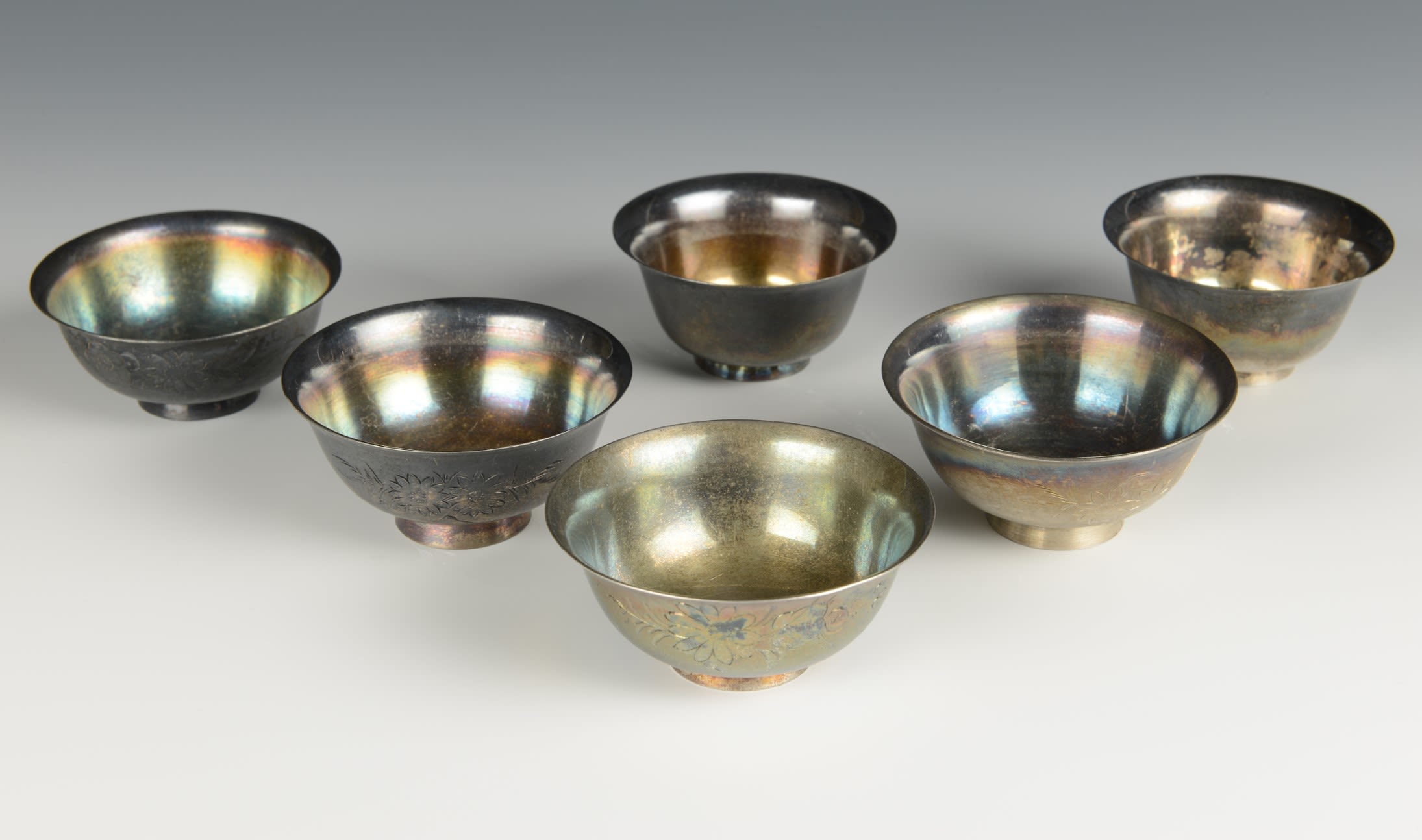 Six small silver cups with wide round tops. The cups are shining in the light and engravings are visible on the sides.