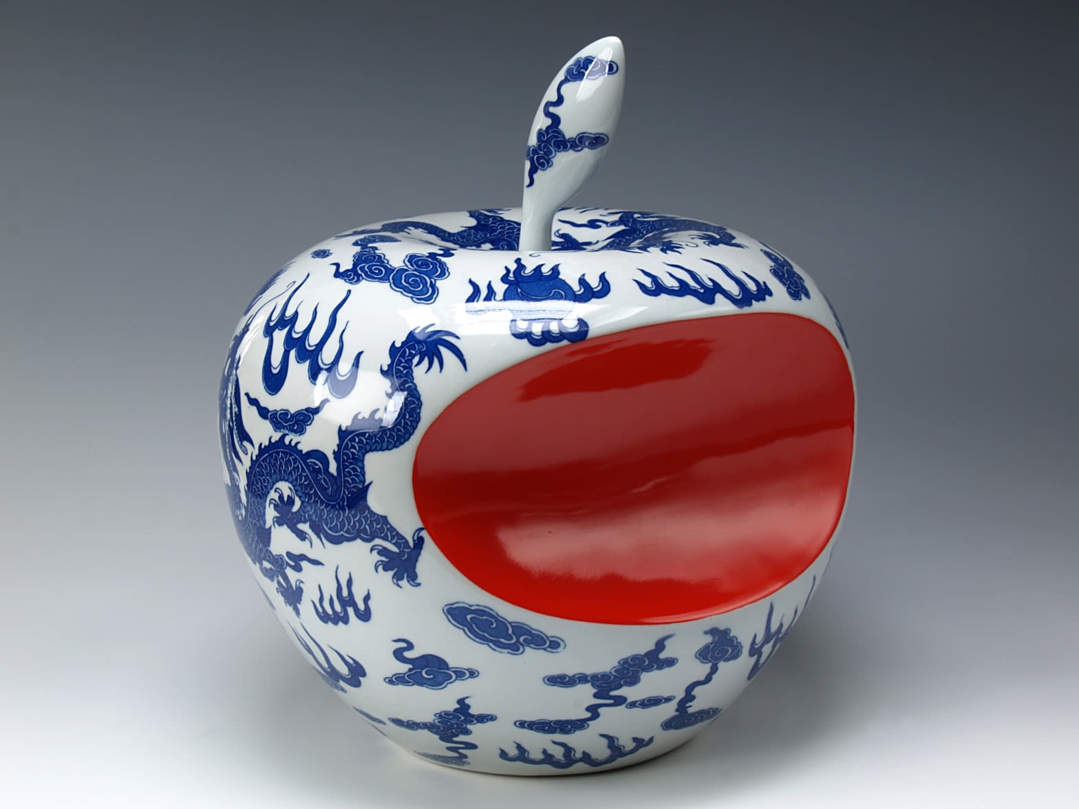 Ceramic sculpture in the shape of an apple with a bite taken from it. The outer visible layer is white with blue patterns in the shape of Chinese dragons. The inner section is bright red.