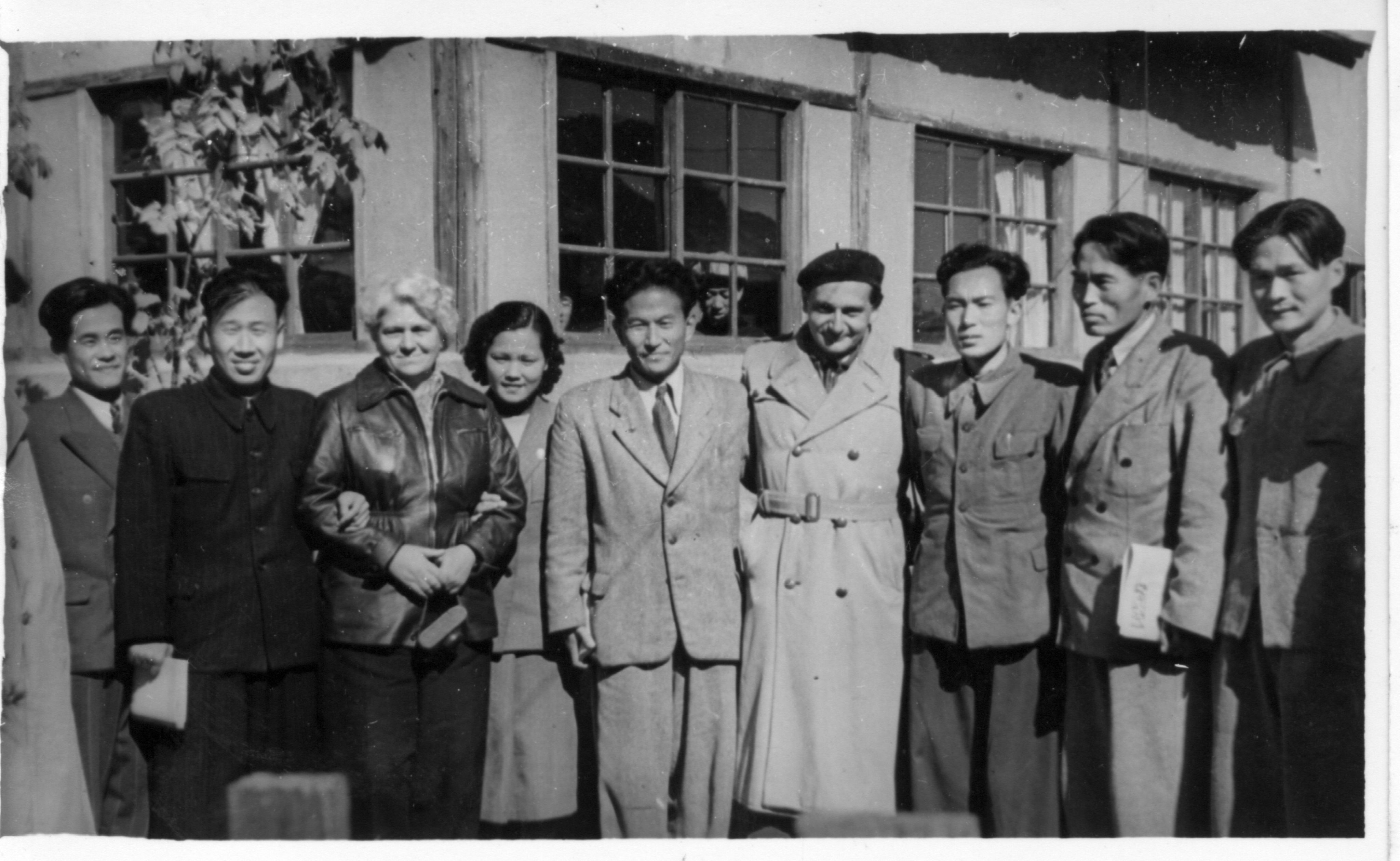 Black and white photograph showing a group of ten people. Erna stands on the left hand side, linking arms with a man dressed in black. She has blonde hair and wears a leather jacket.