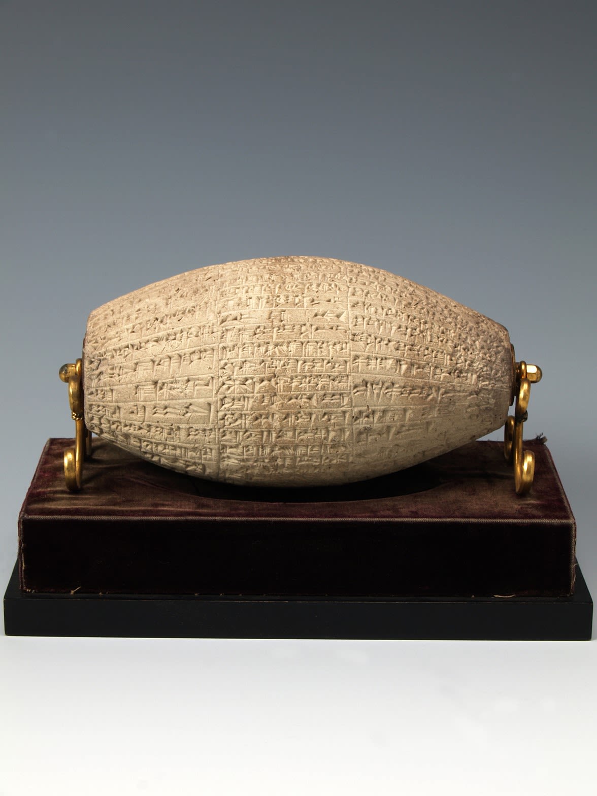 A beige clay cylinder in a horizontal tubular shape. The cylinder is supported by gold coloured clasps on the right and left and sits on a wooden base