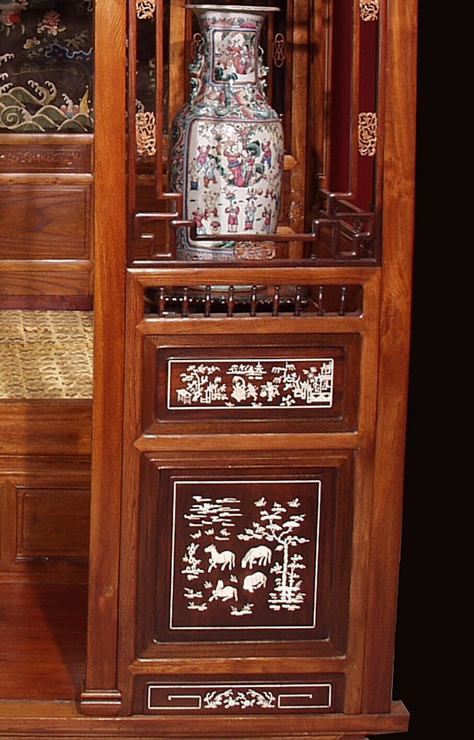 an angled view of the Chinese bed, inside the wooden frame is a yellow blanket and a backdrop showing waves and flowers. There is also a vase within the wooden frame