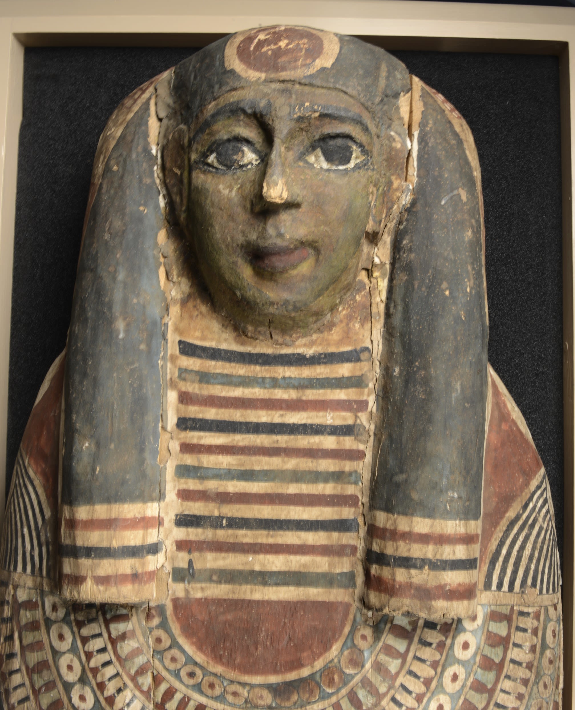 Close up on the face of a Mummy coffin. The depicted figure has large eyes and a headpiece which covers the left and right side of the head.