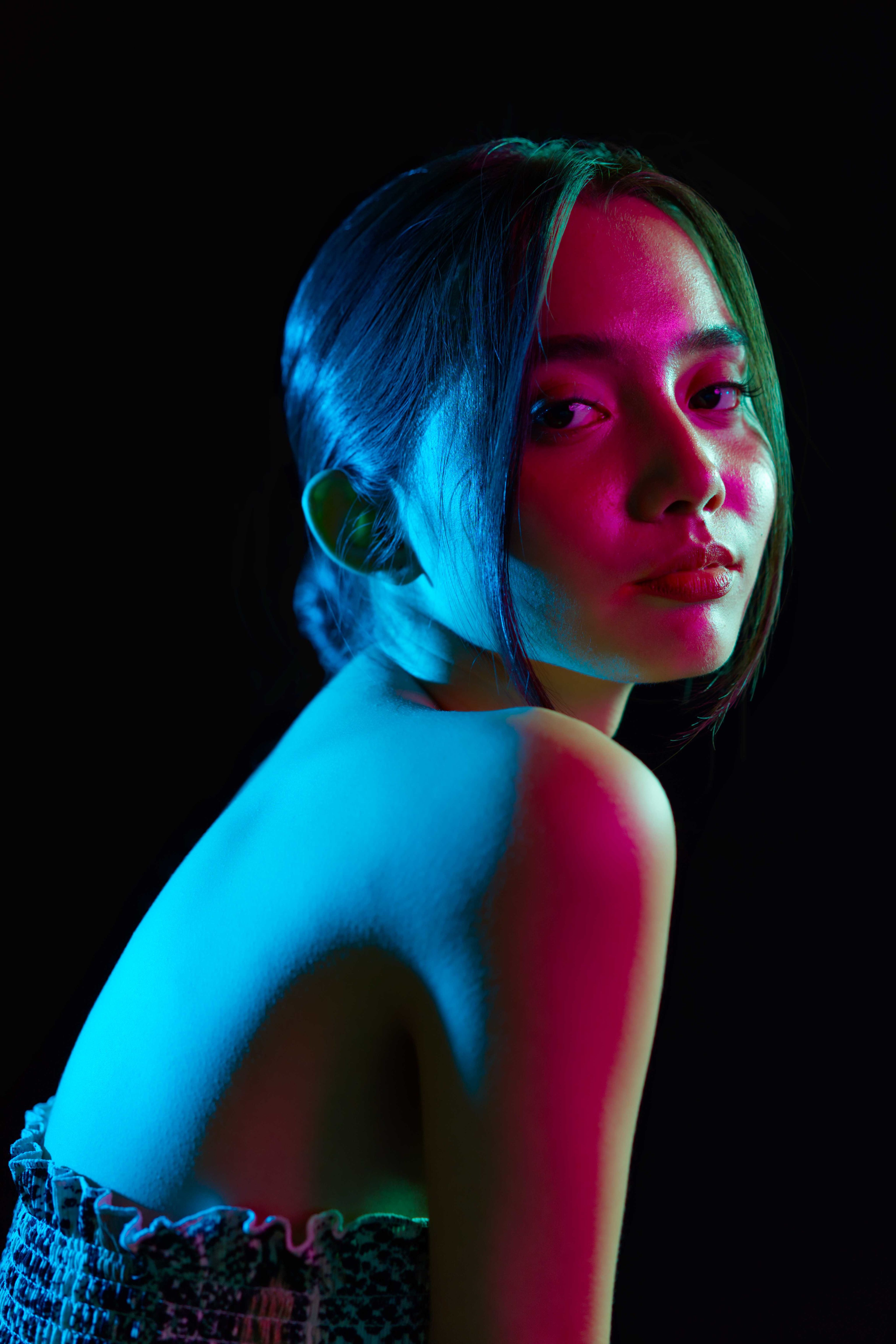 vDigital photography of a person sat with their body facing right, but their face turned to the camera. On their exposed back the lighting is blue, on their face the lighting is red. 