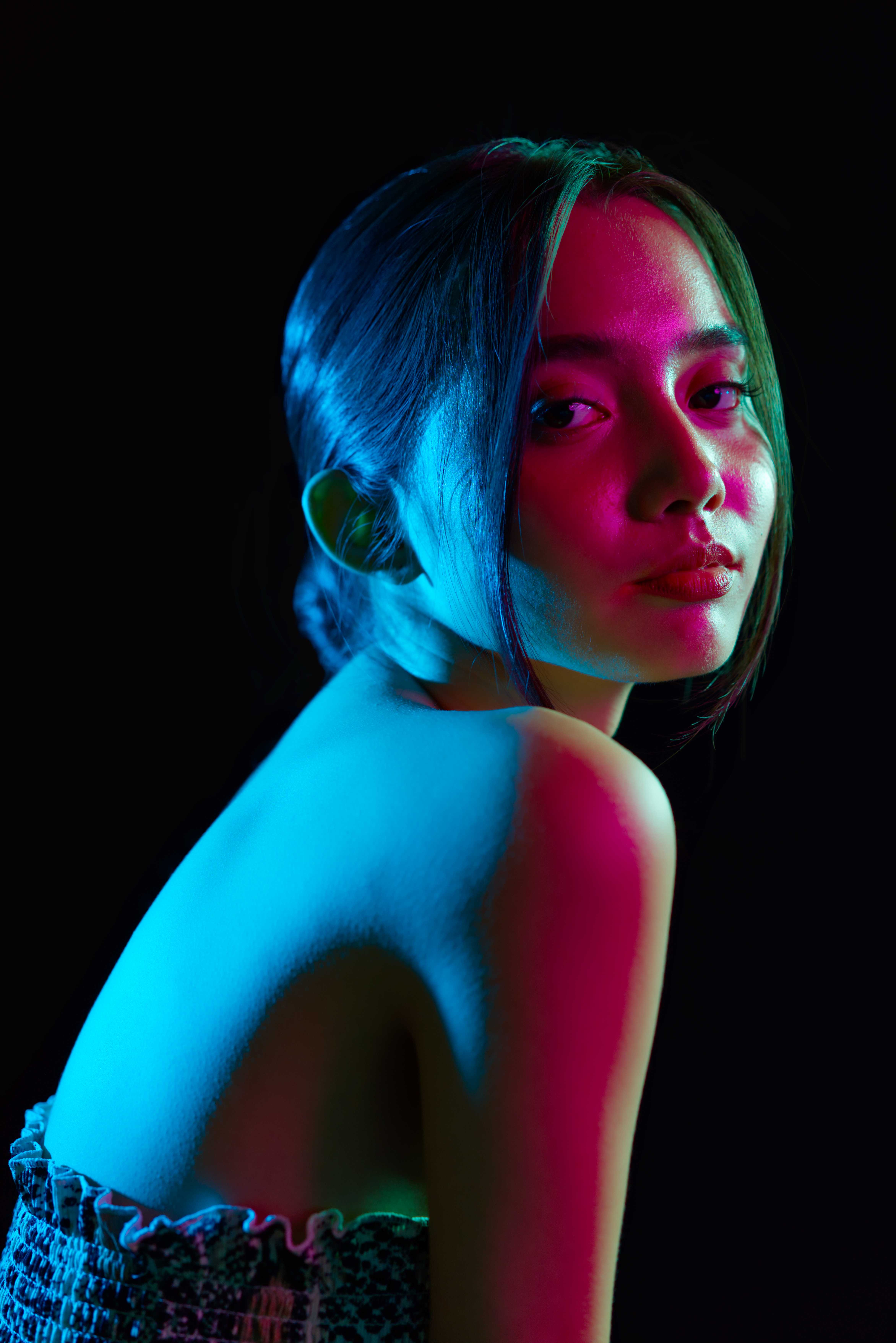 Digital photography of a person sat with their body facing right, but their face turned to the camera. On their exposed back the lighting is blue, on their face the lighting is red.