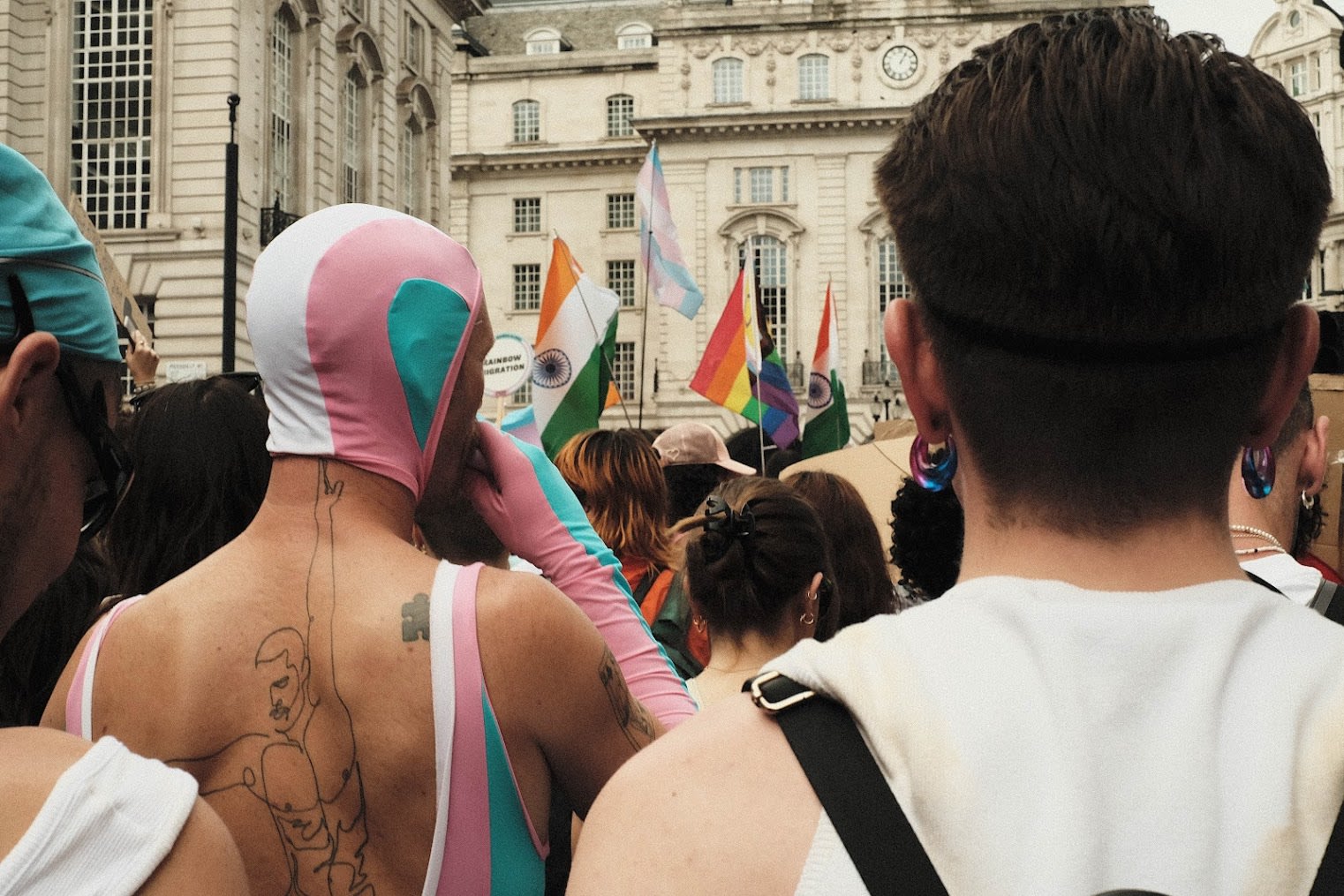 A colour photograph of a pride parade showing the backs of peoples heads. we see rainbow flags and a person dressed in the trans flag colours.