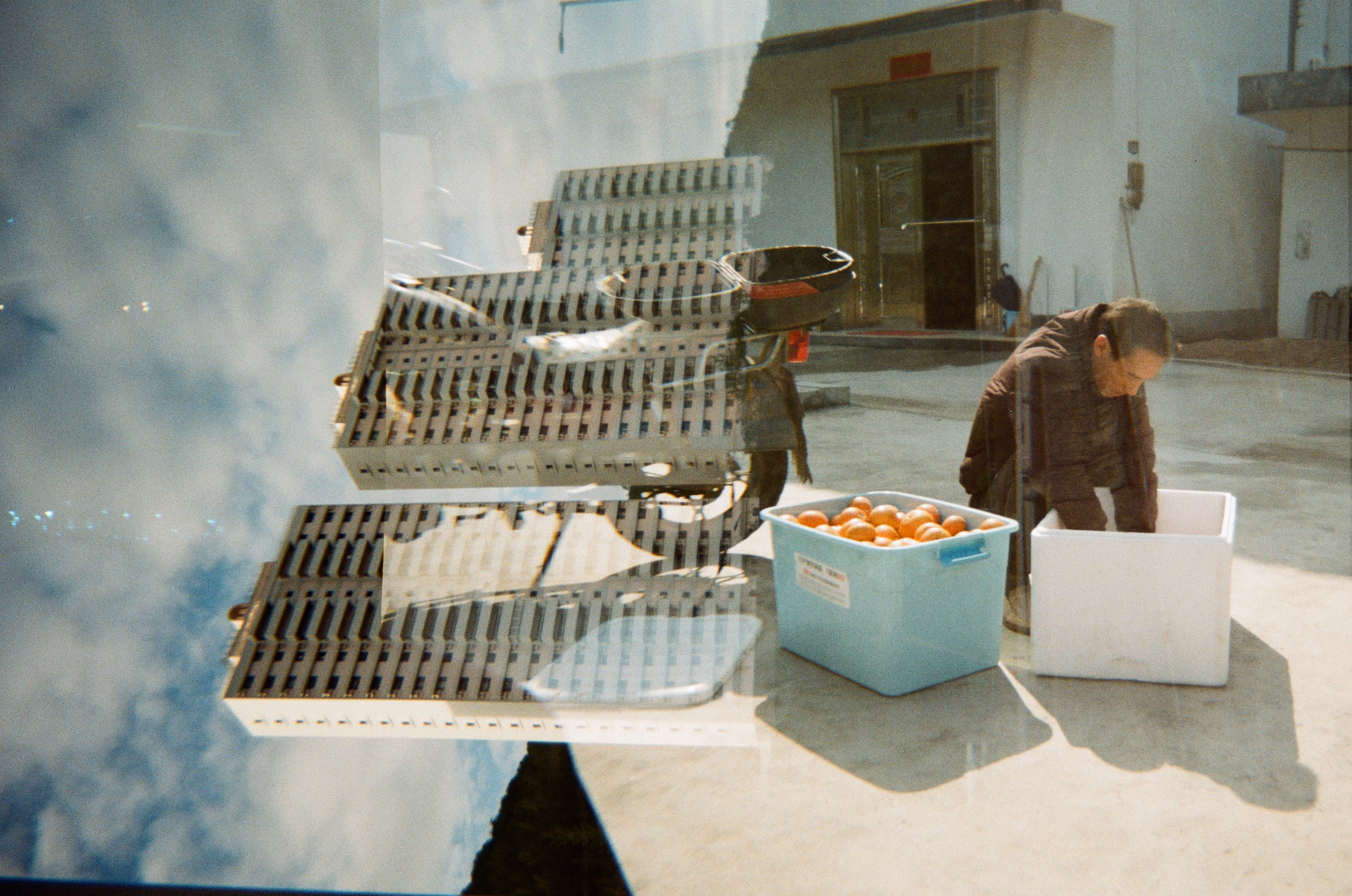 Double exposure colour photograph of a set of high rise buildings and a person next to a motor bike in the road crouching down with boxes of oranges.