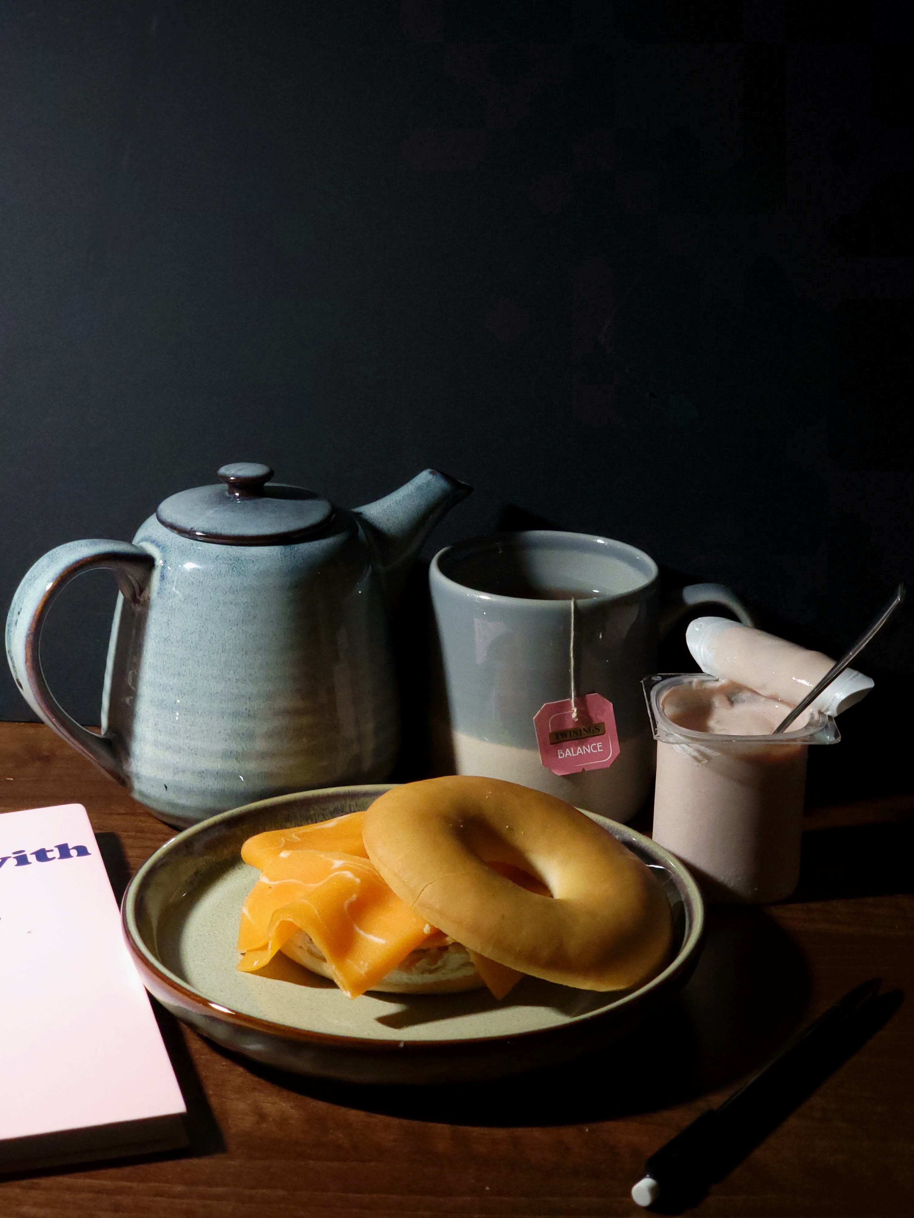 Colour photograph of small tea pot, mug with tea bag in, open full yogurt with spoon and filled bagel on a plate.