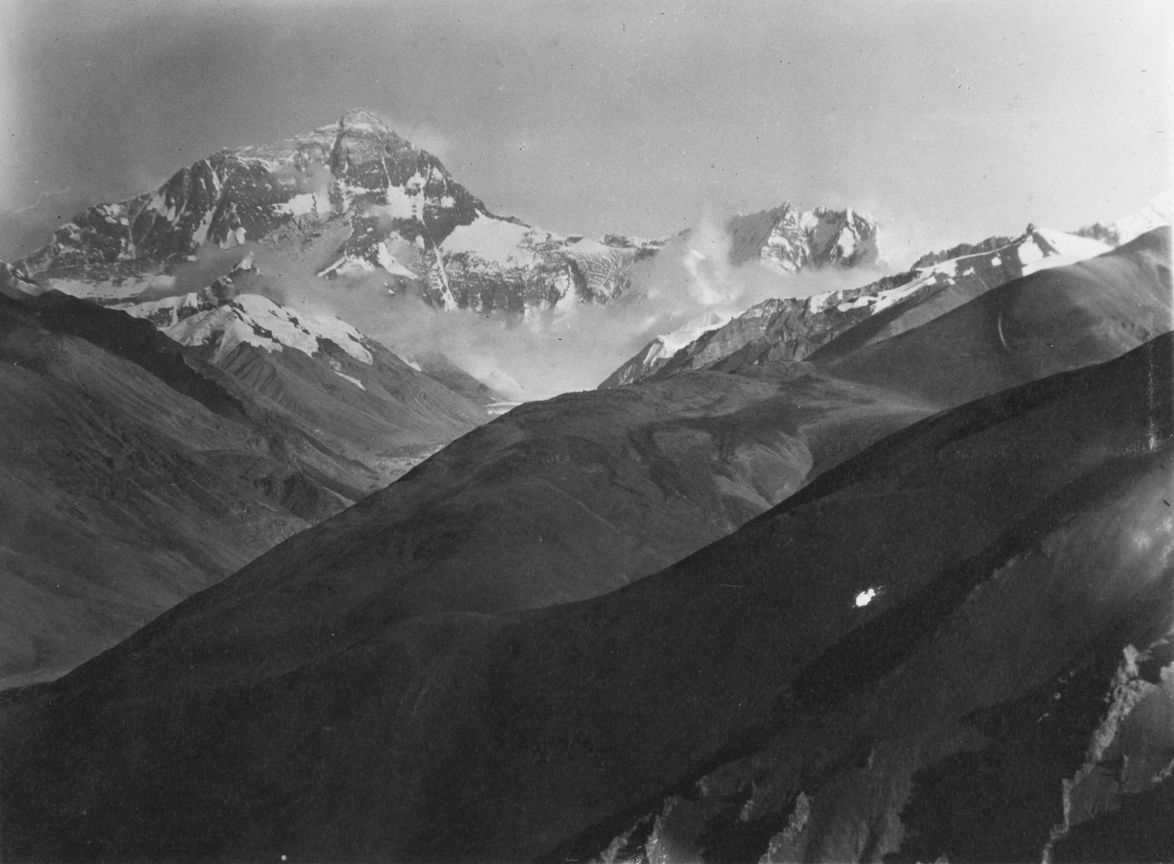 Black and white photo of a large mountain range. A tall snow covered mountain appears in the distance emerging from the clouds. 