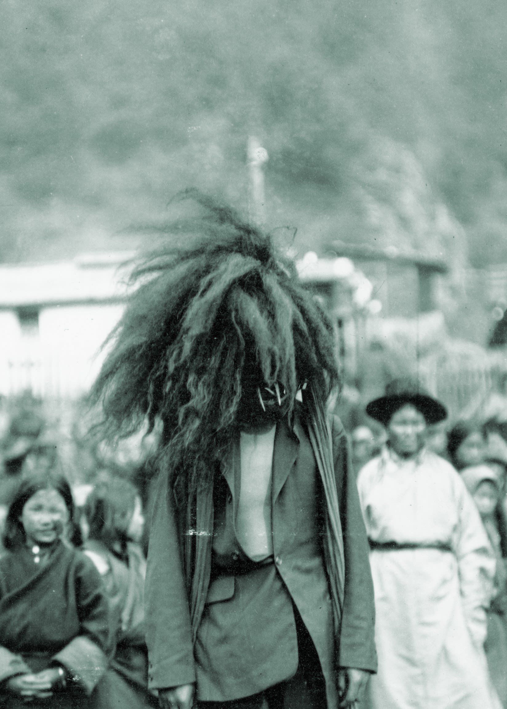Black and white photo of a person wearing a mask and long coat, standing outdoors with blurred figures and trees in the background