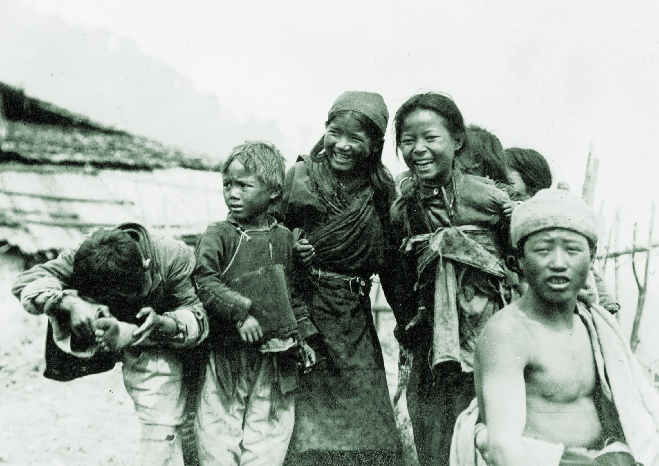 A greyscale photo with a green tint of seven children, in order from the left to the right of the image, the first child is hiding his face whilst holding his foot, the next child is looking to the left, the next two children are laughing and bend towards each other. The two children towards the back right have their backs turned and the child in the front right has a startled expression.