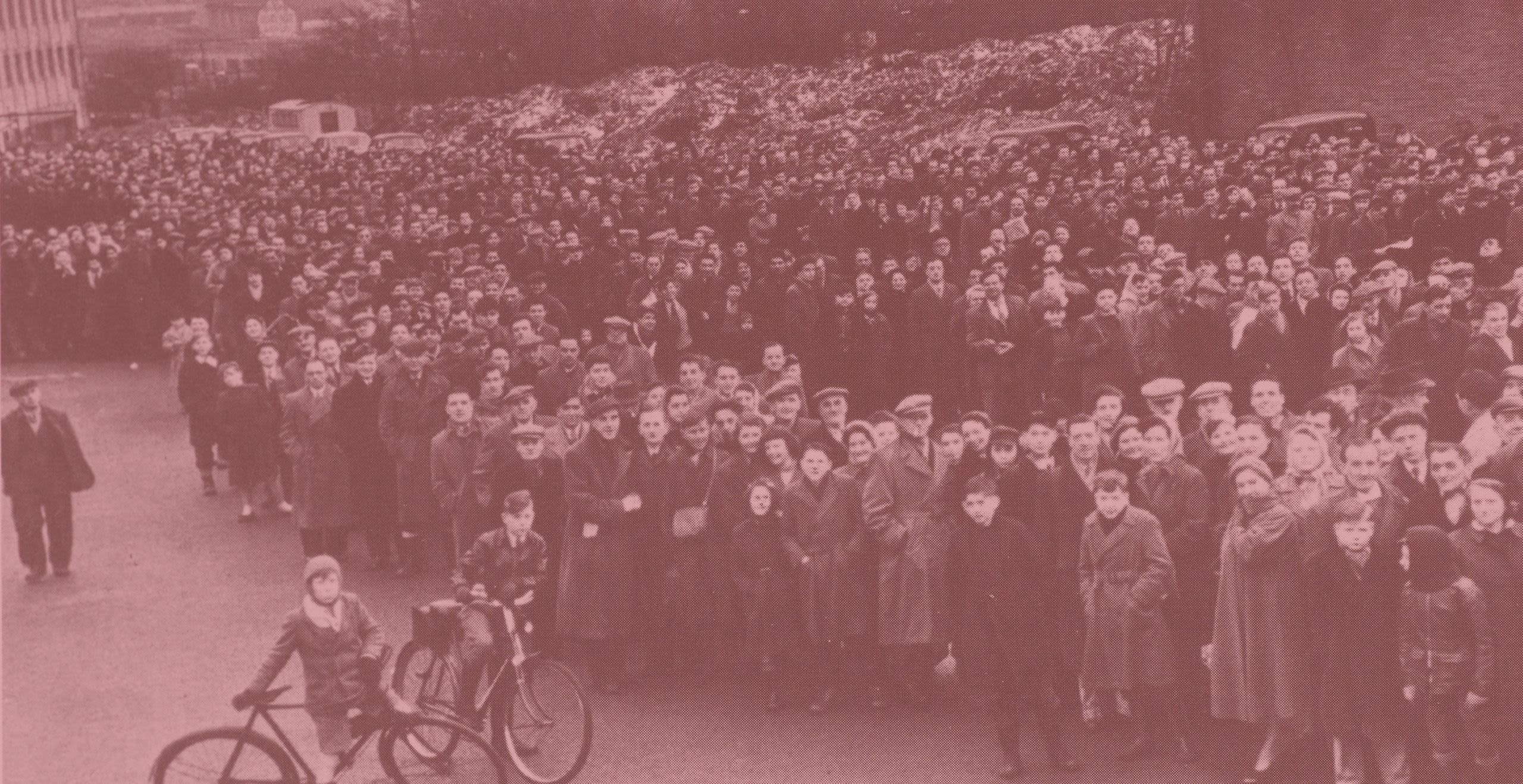 Black and white photo of a long line of people dressed in 1950s long coats and flat caps.