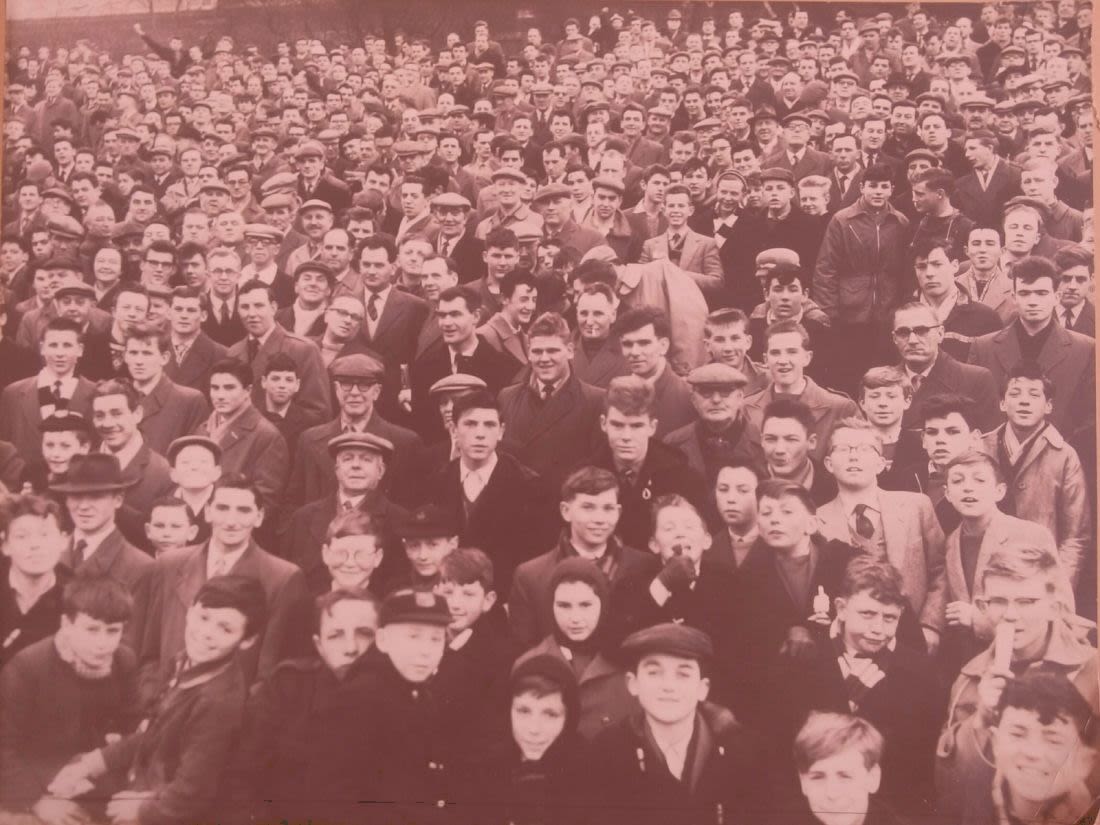 Photo of a crowd of football fans in the 1950s. The crowd is packed with men in coats and caps. One woman, wearing a hood to cover her hair is visible in the centre of the image.