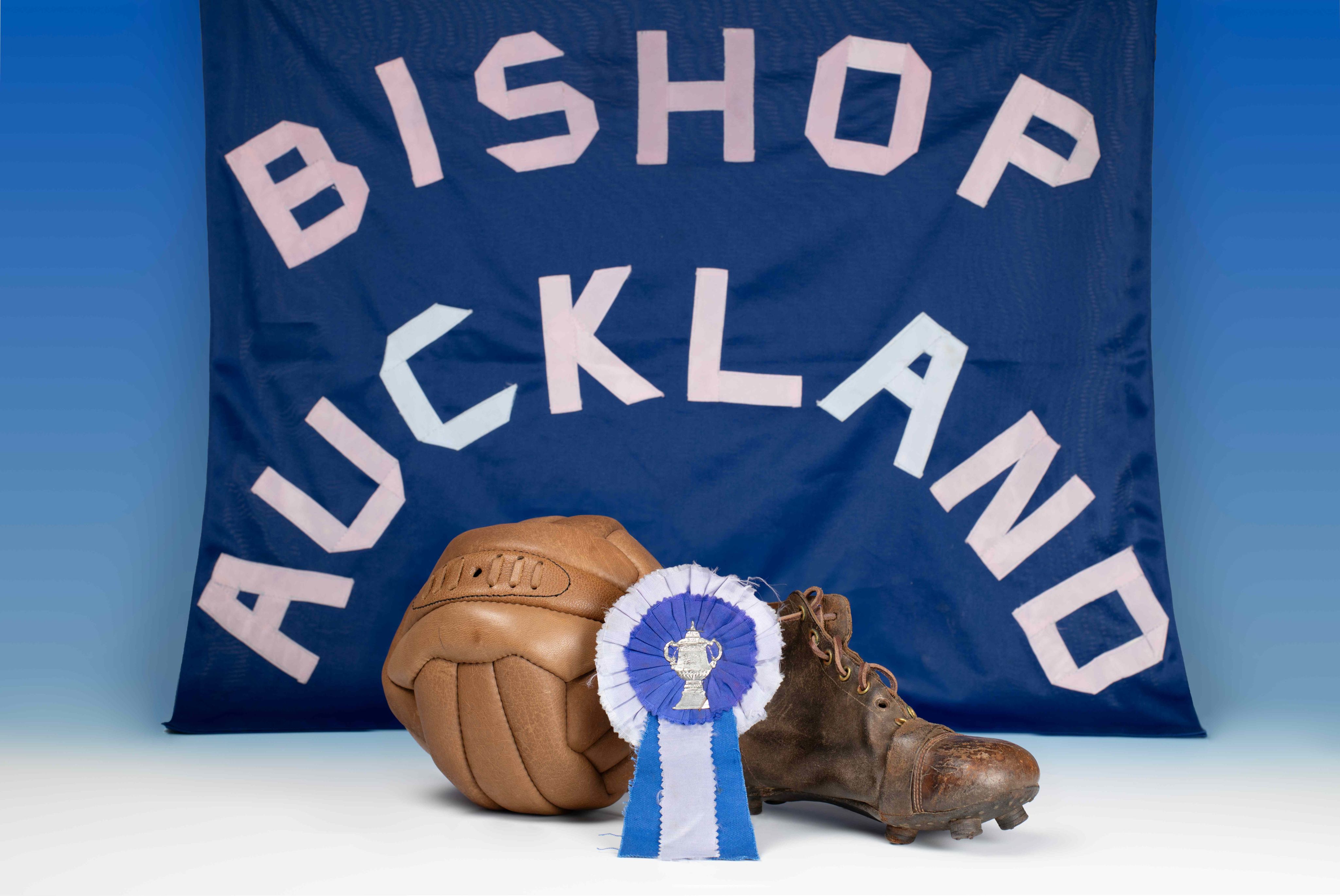 A blue banner reading Bishop Auckland behind a 1950s football boot and ball and a blue and white rosette showing the FA Cup.