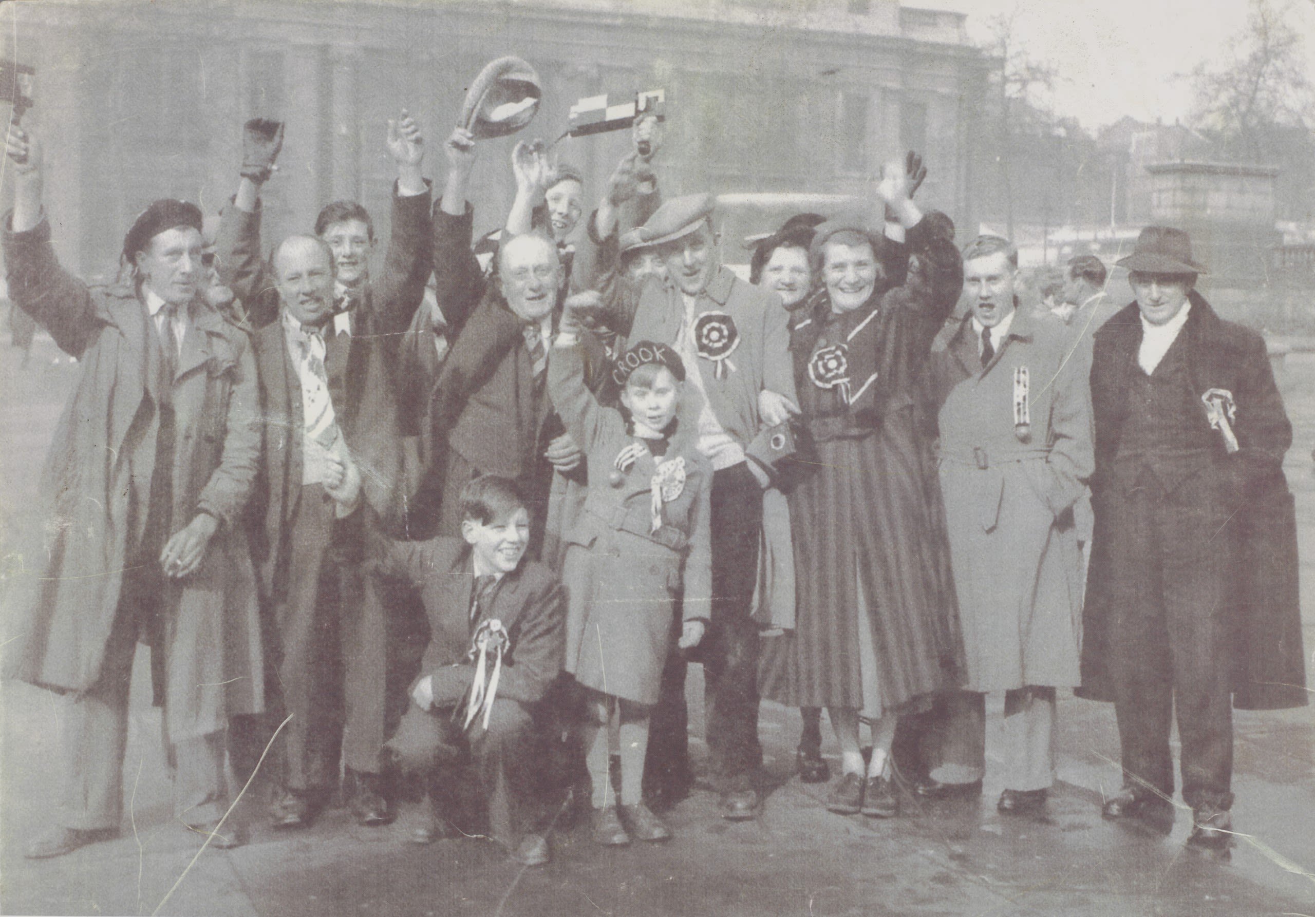 Black and white photo of a group of celebrating football supporters, including men, women and children, dressed in 1960s clothes, wearing rosettes and waving rattles.