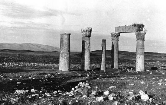 Black and white photograph, outdoors, of moorland and collapsed stone buildings with only columns remaining.