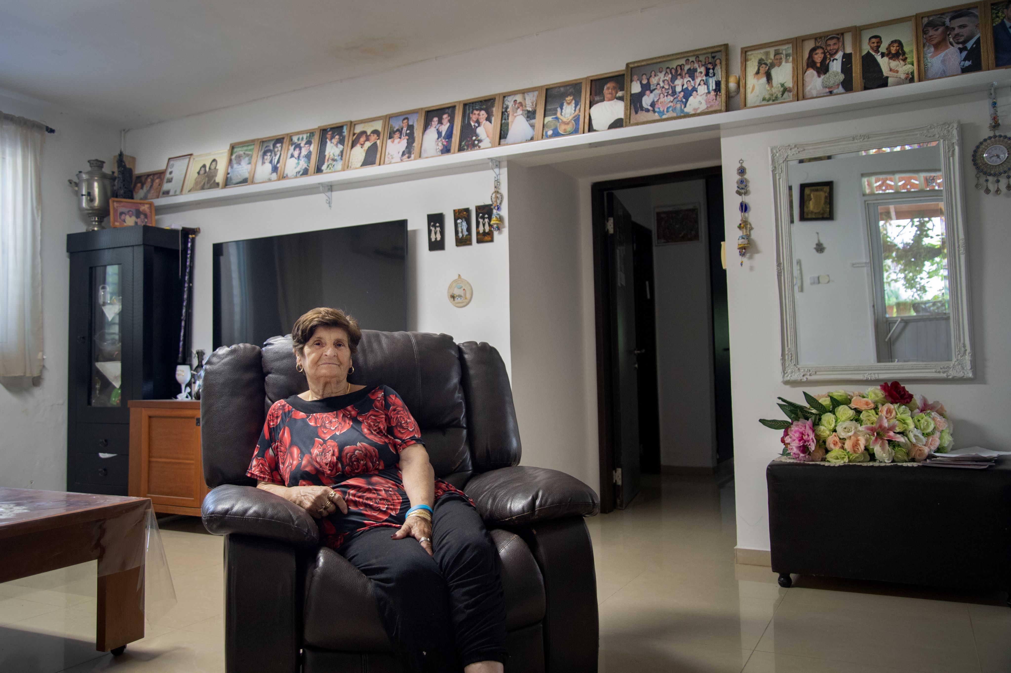 Colour photograph, indoors, woman seated on brown sofa looking towards camera, in the background is a row of family photographs, furniture and decorative wall hangings such as the Devil's Eye.