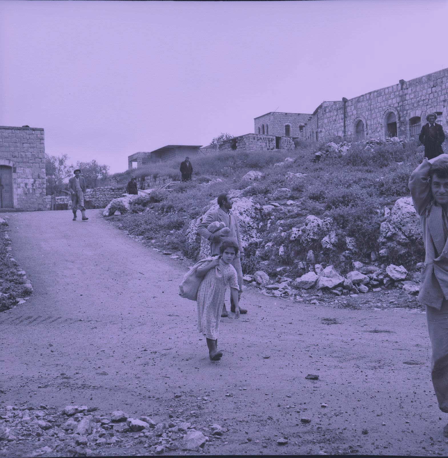 Black and white photograph in purple tones, outdoors, man and woman carrying luggage in close up, stone house and three other people in distance.