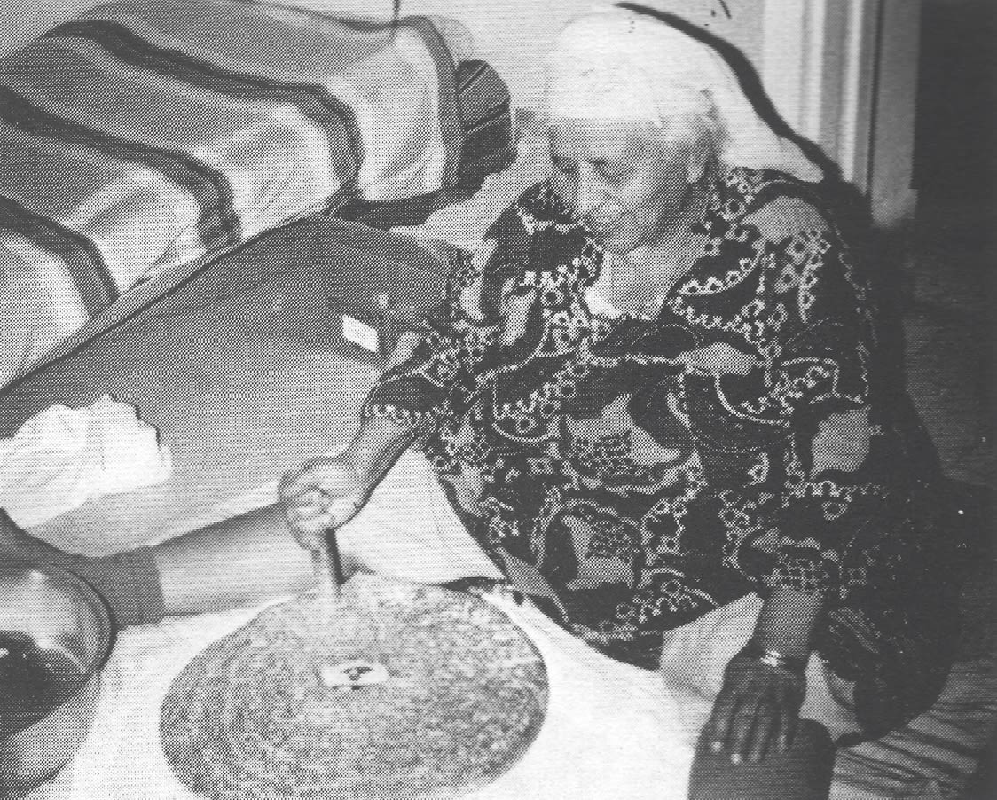 Black and white photograph, indoors, of a woman sitting on the floor with her legs spread out and a large piece of pie in front of her.
