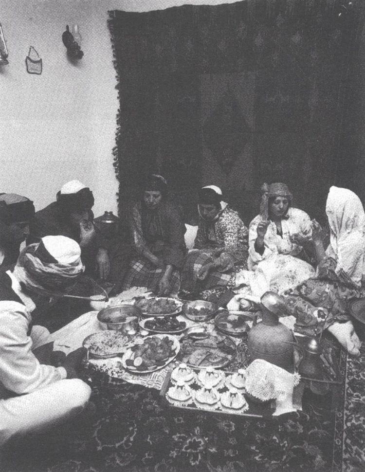 Black and white photograph, indoors, a group of people sitting at a low table filled with food.