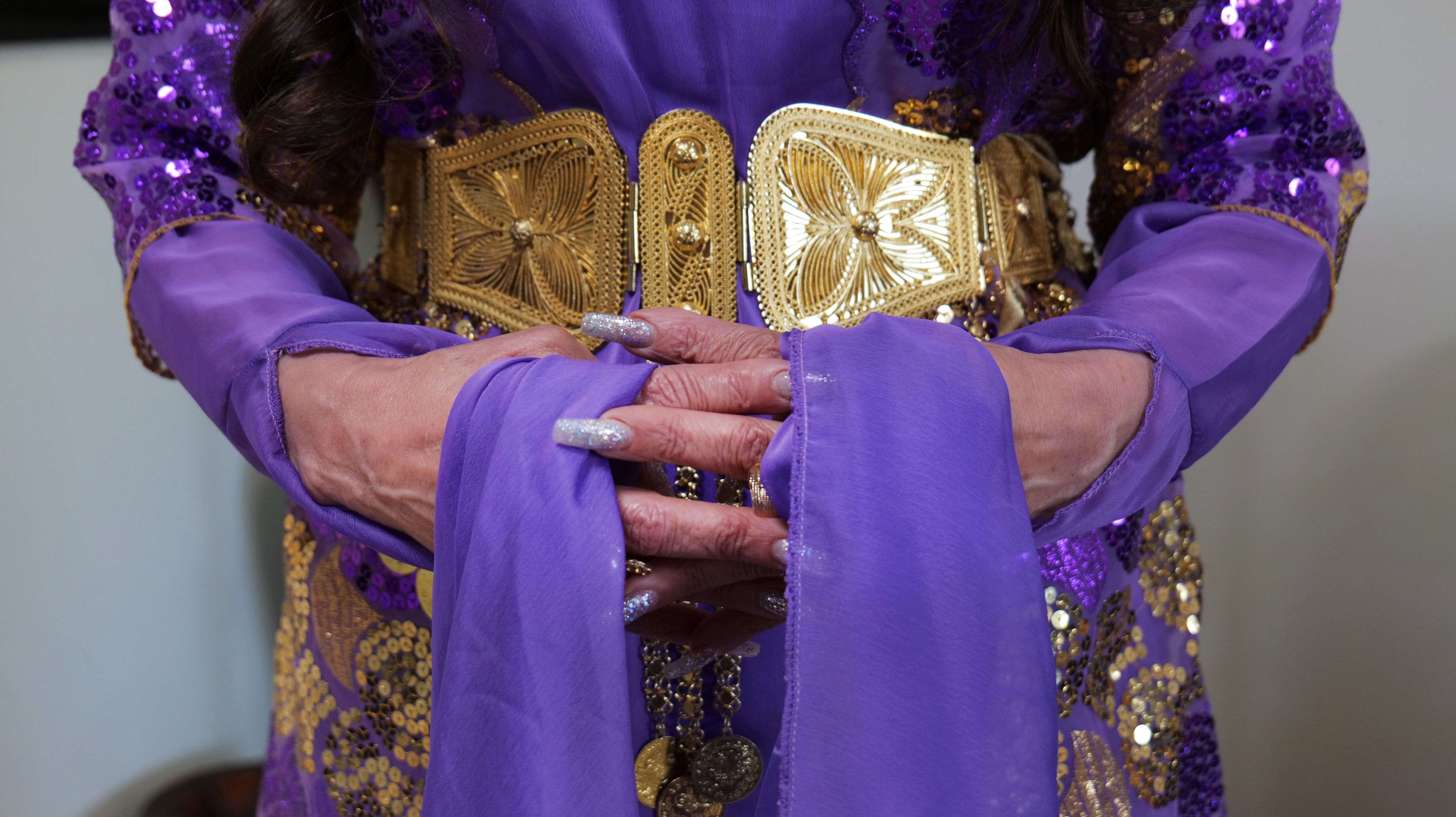 Colour photograph, close up, woman's folded hands, purple dress with gold sash and trim.
