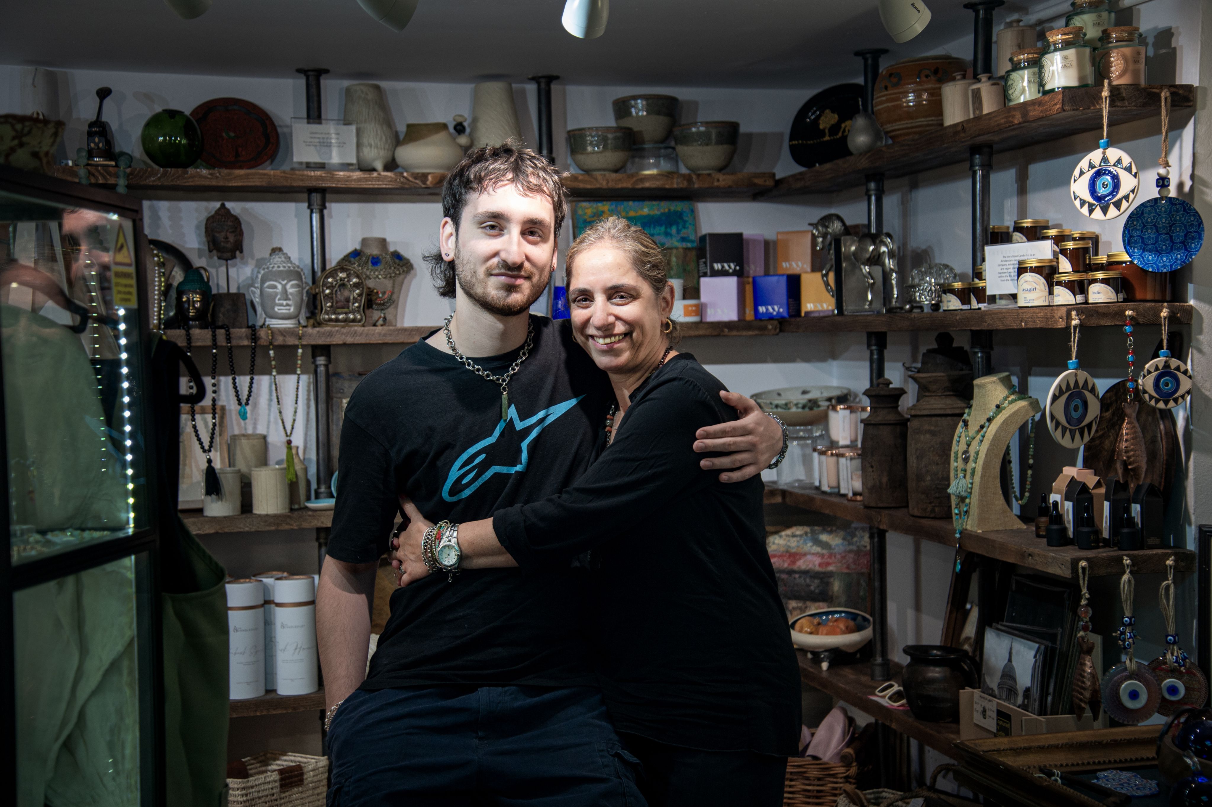 Colour photograph, indoors, man and woman look towards camera and embrace each other, shelves full of things in background.