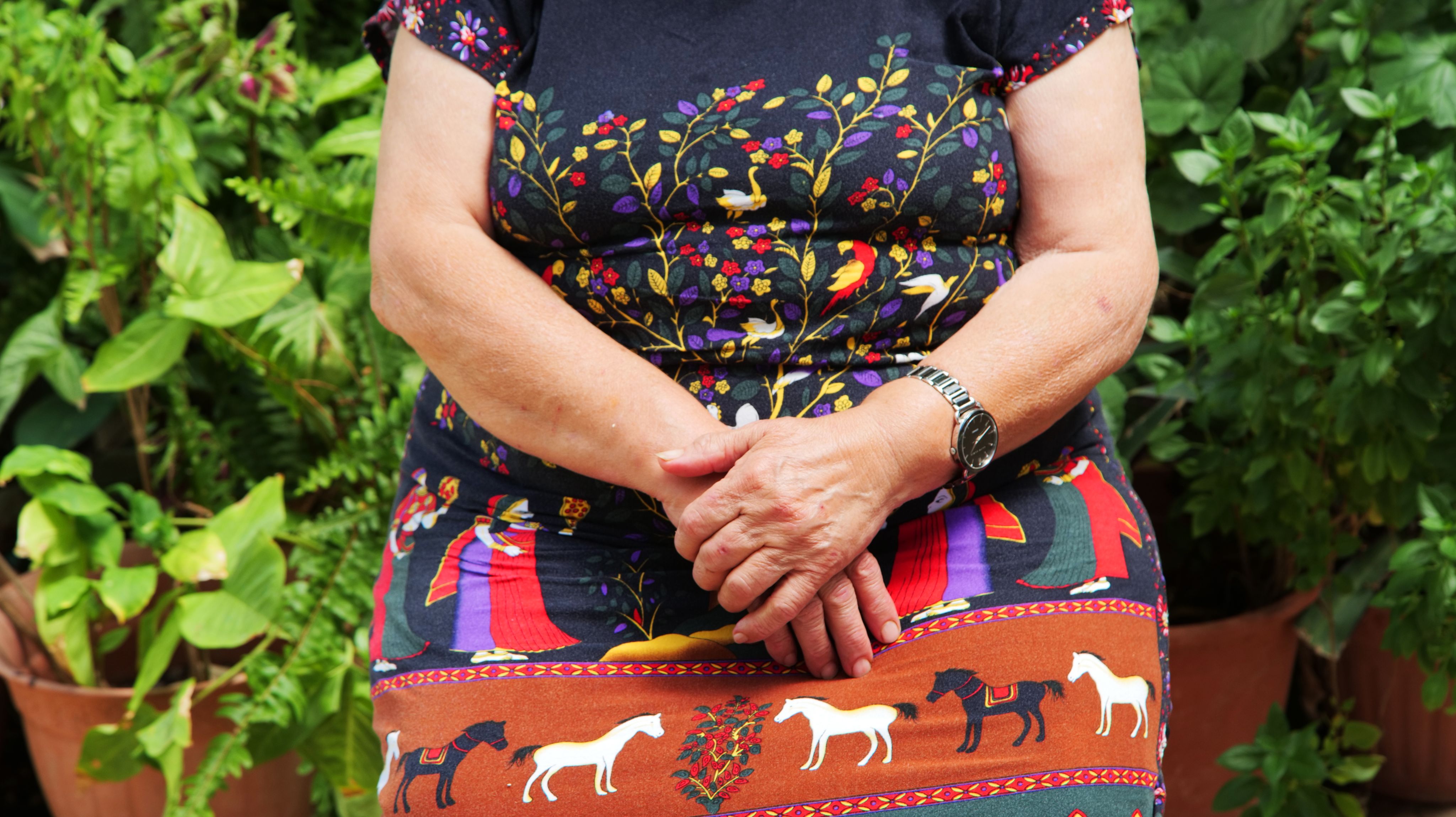 Colour photograph, close up, of woman's folded hands, with trees, birds, people and horses on her dress.