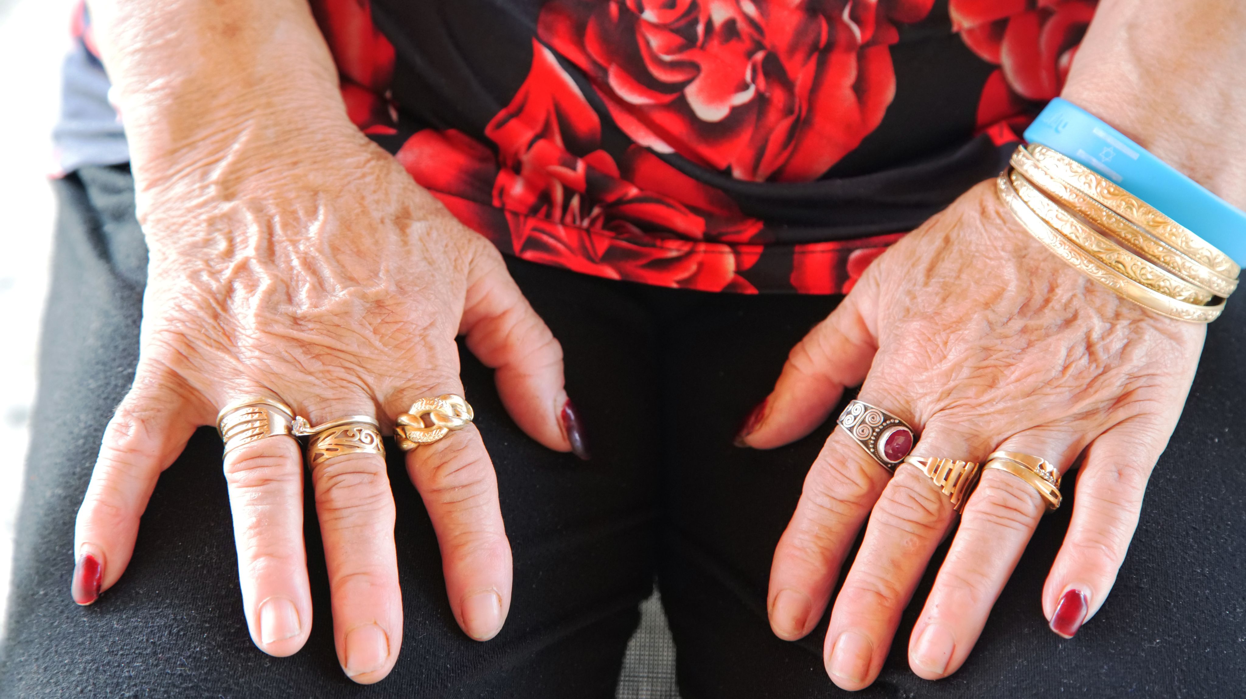 Colour photograph, close up, of woman's folded hands, red flowers on dress, many rings on hands.