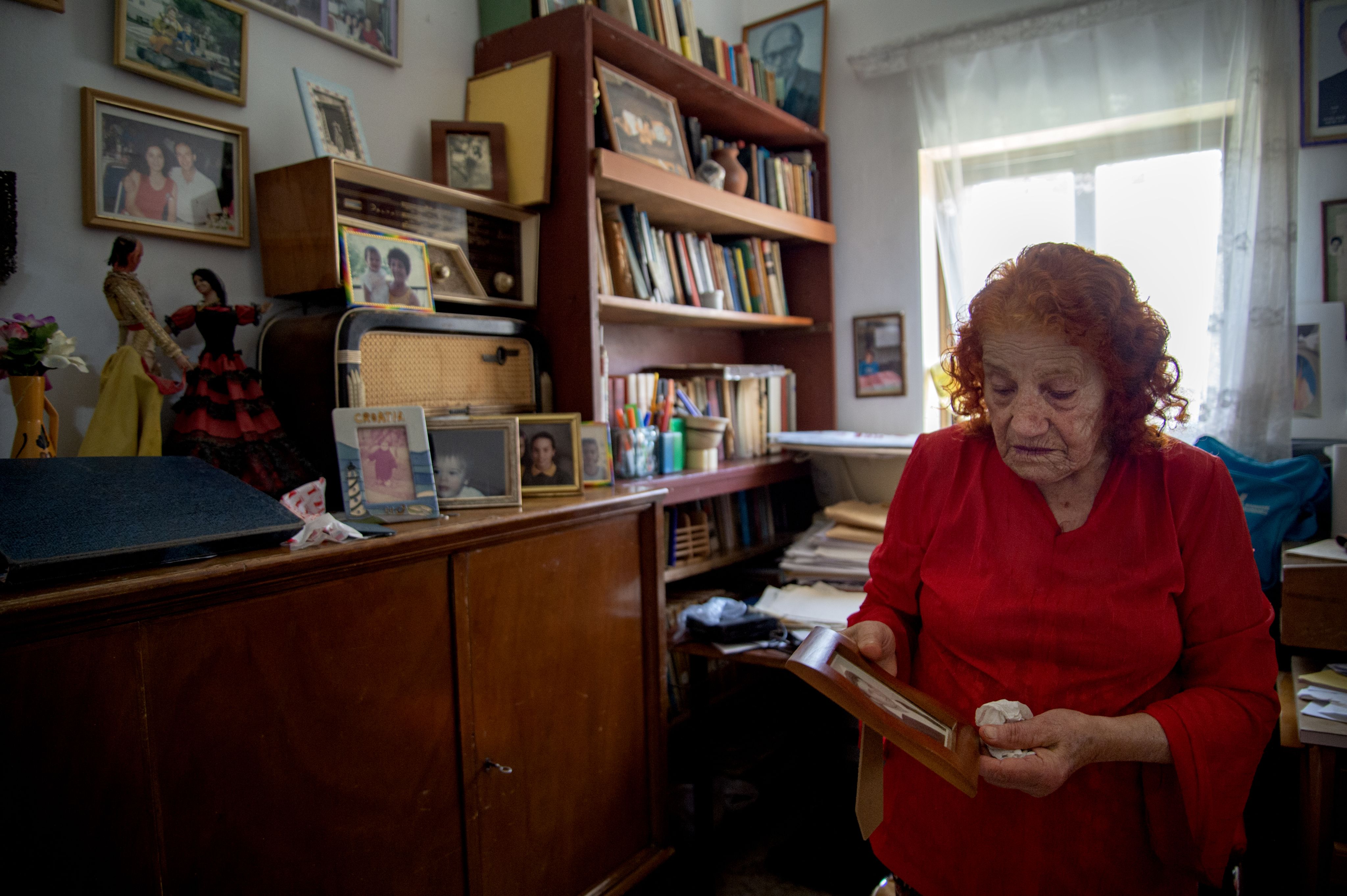 Colour photograph, indoors, woman looking at a black and white photograph, cupboard full of photographs and books.