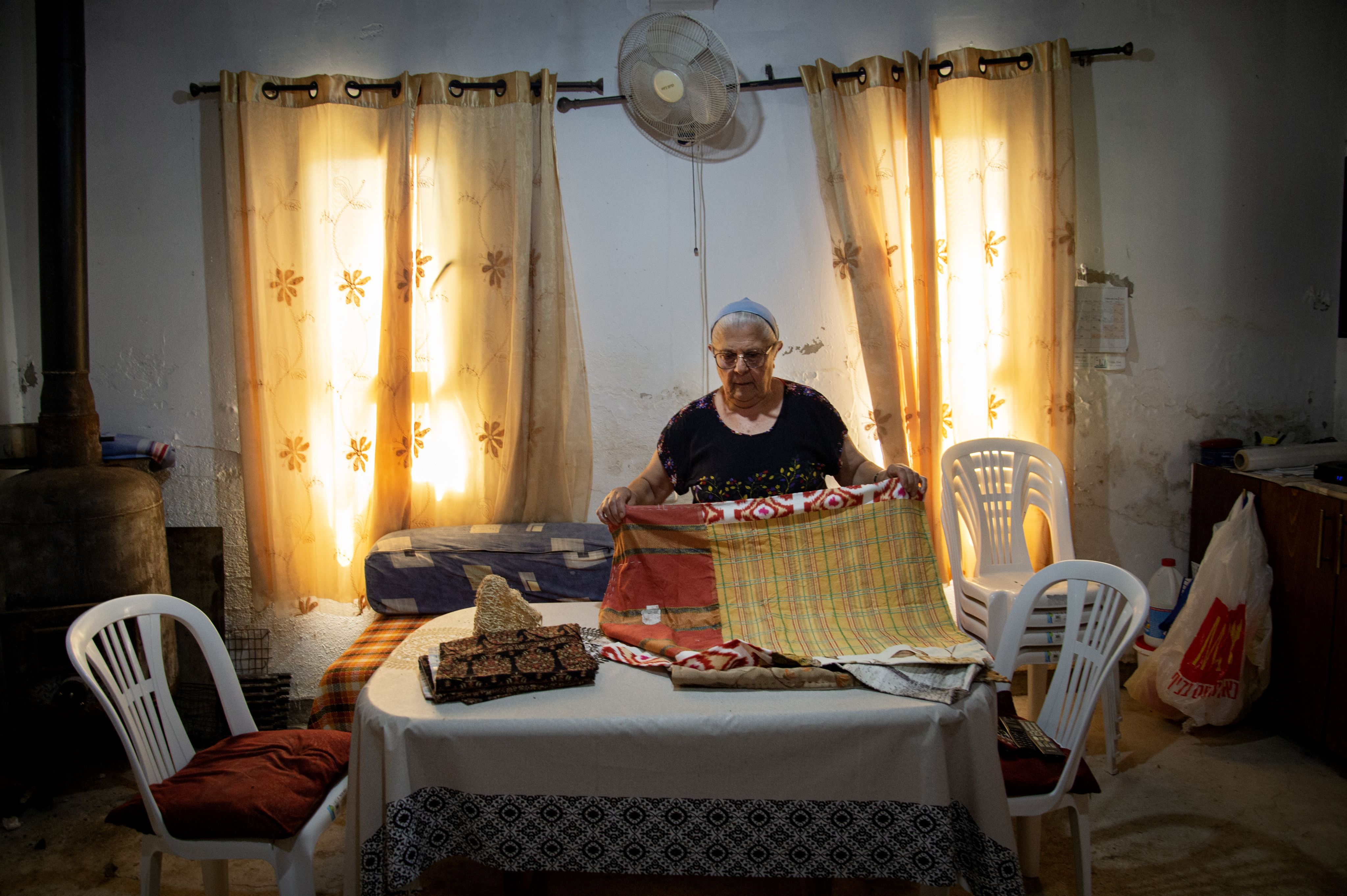 Colour photograph, indoors, woman folding a piece of fabric, curtains in the background, warm white sunlight coming through the window.