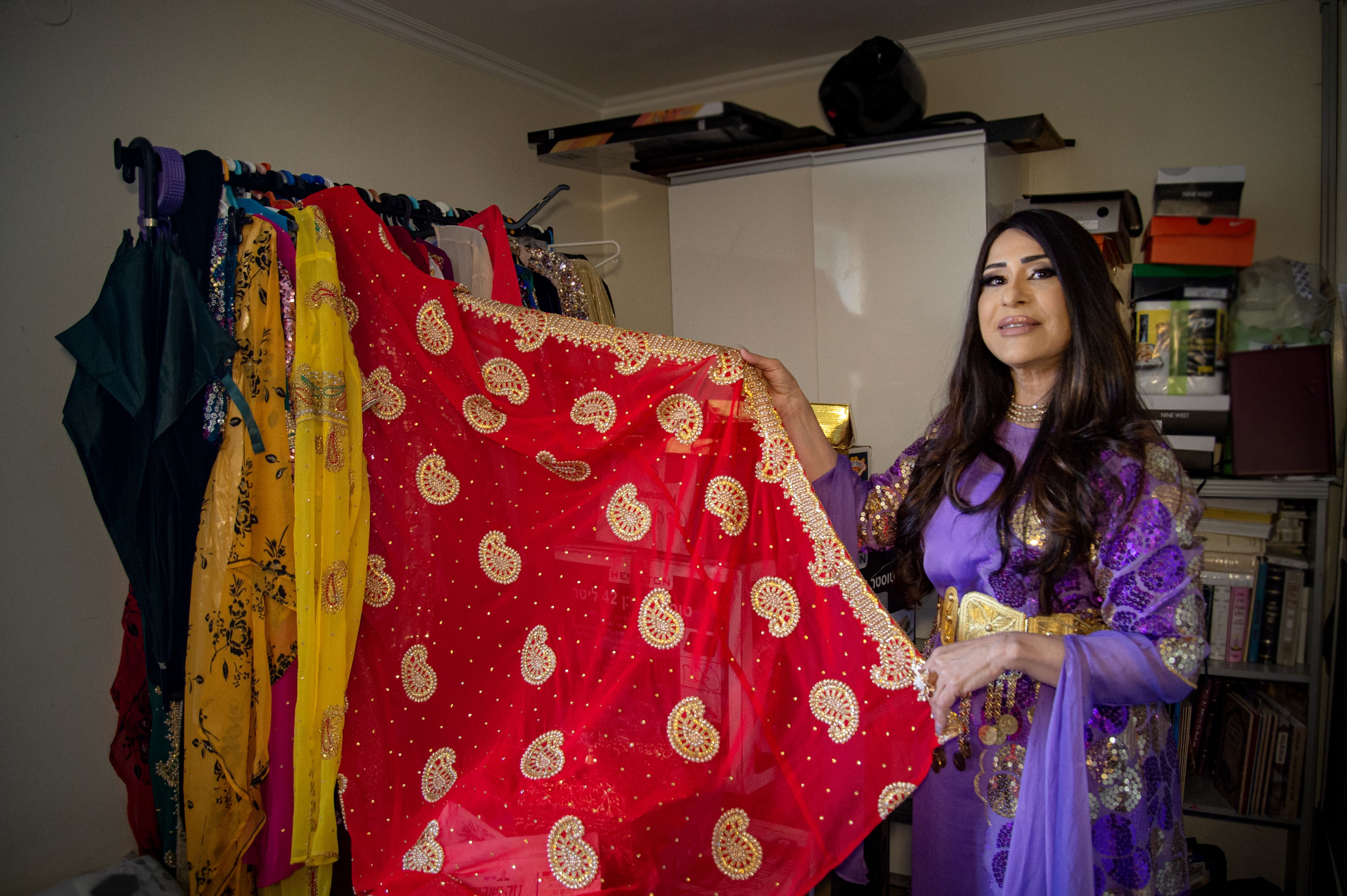 Colour photograph, indoors, woman in purple dress showing red textile, clutter and books in background.