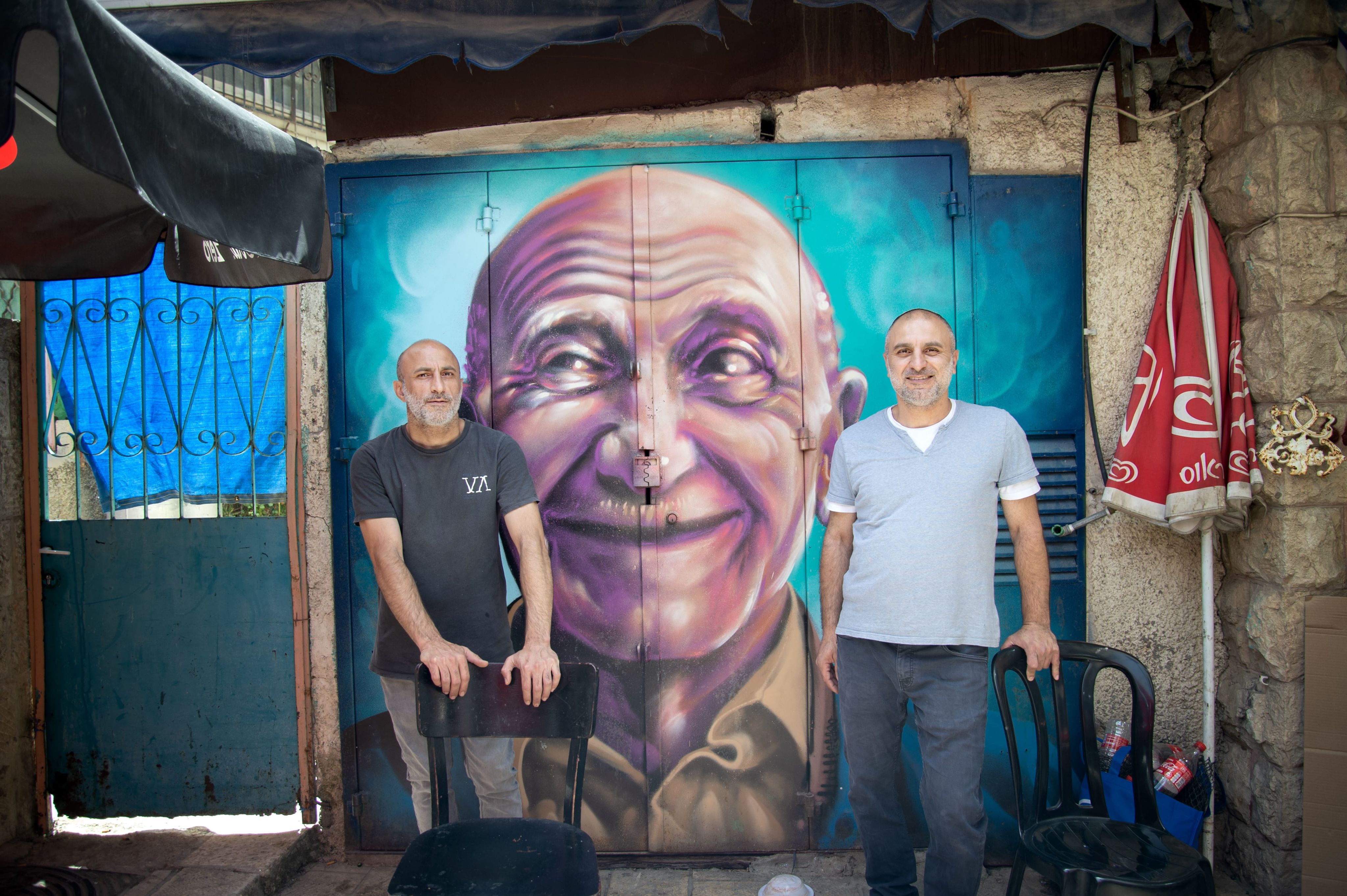 Two men stand in a casual patio seating area of a restaurant in front of a large painted mural of a bald man.