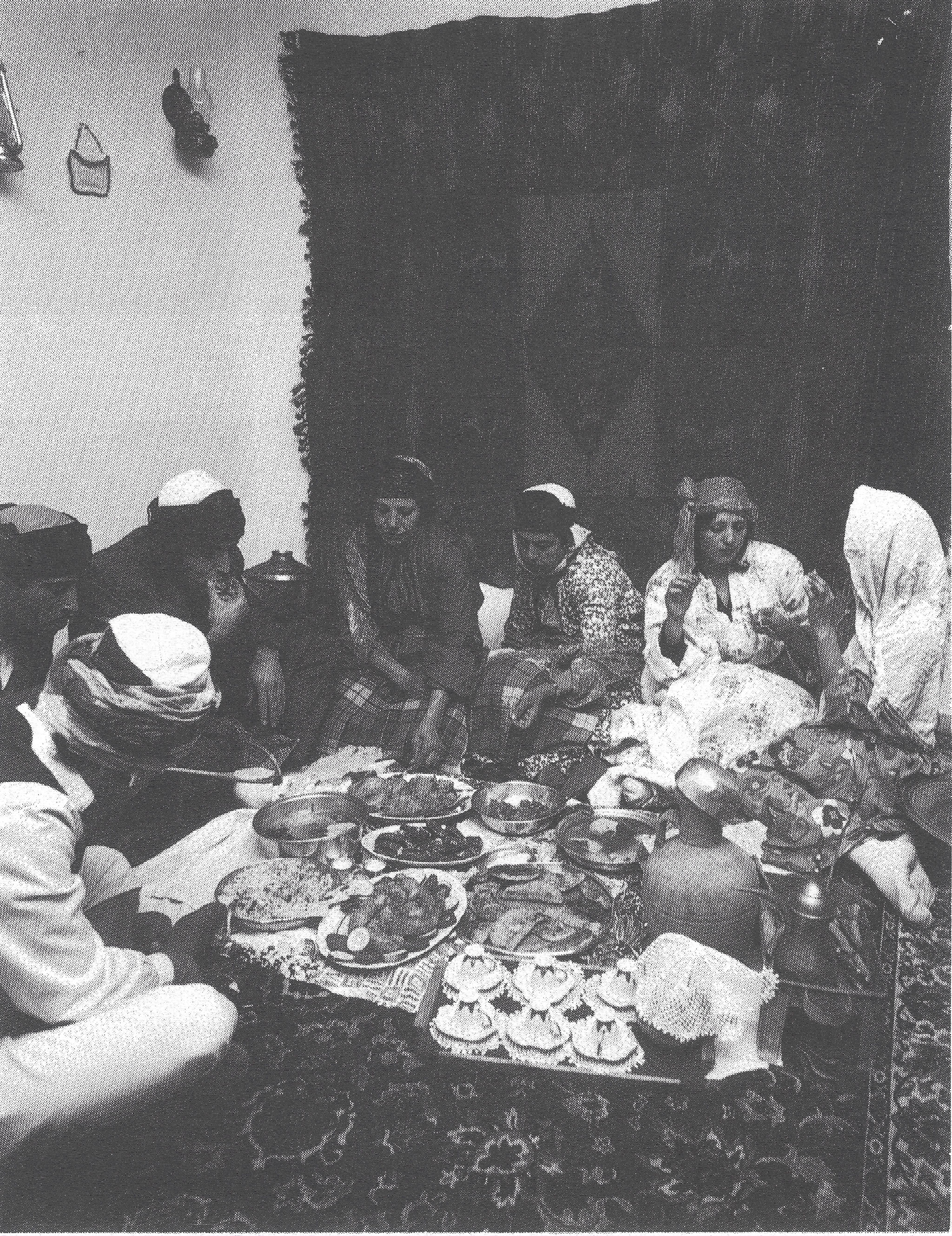 Black and white photograph, indoors, a group of people sitting at a low table filled with food.