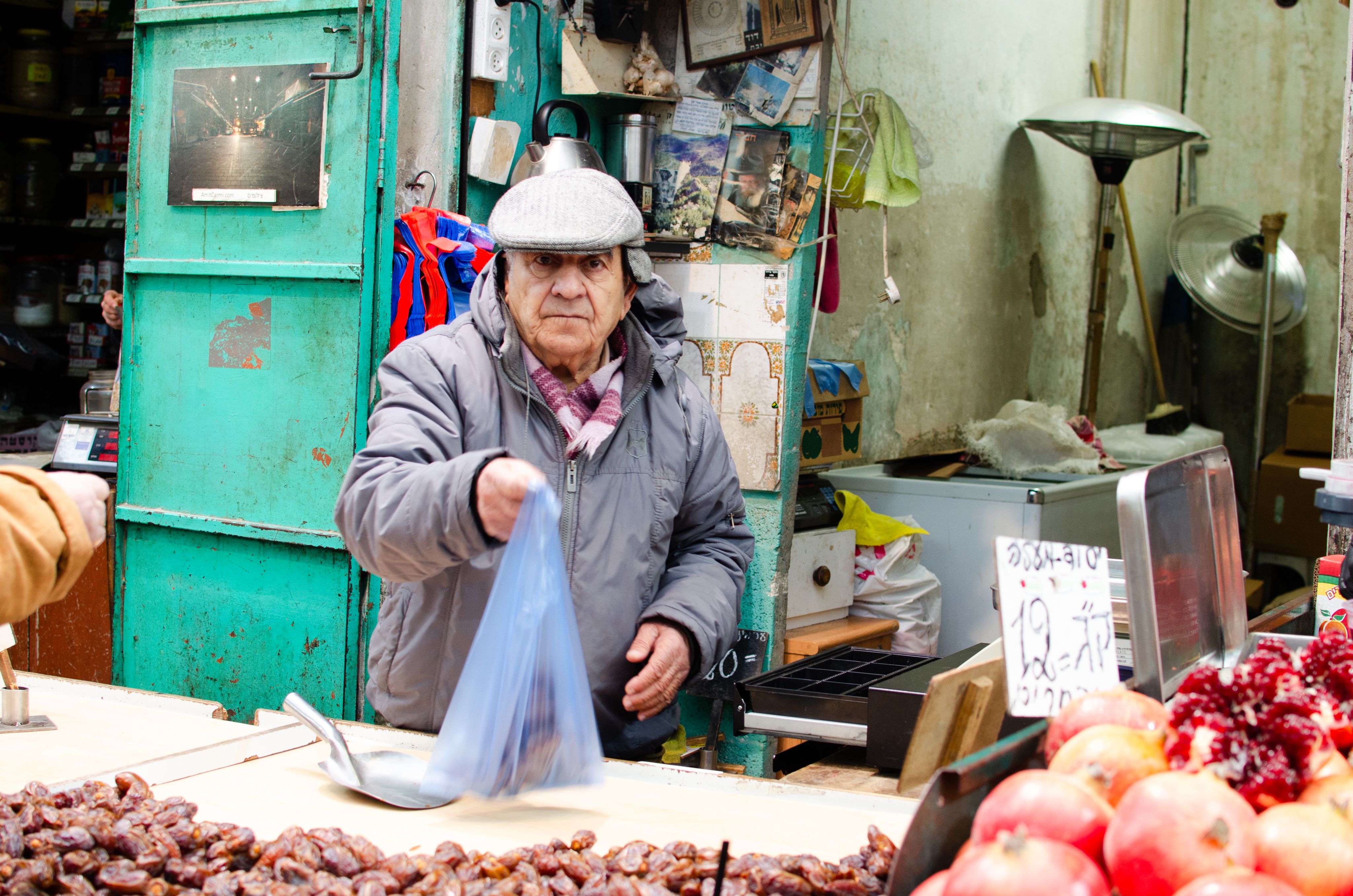 Colour photograph, outdoors, man hands bag of dates to camera, dates and pomegranates in close up, green door, various posters and clutter in background.