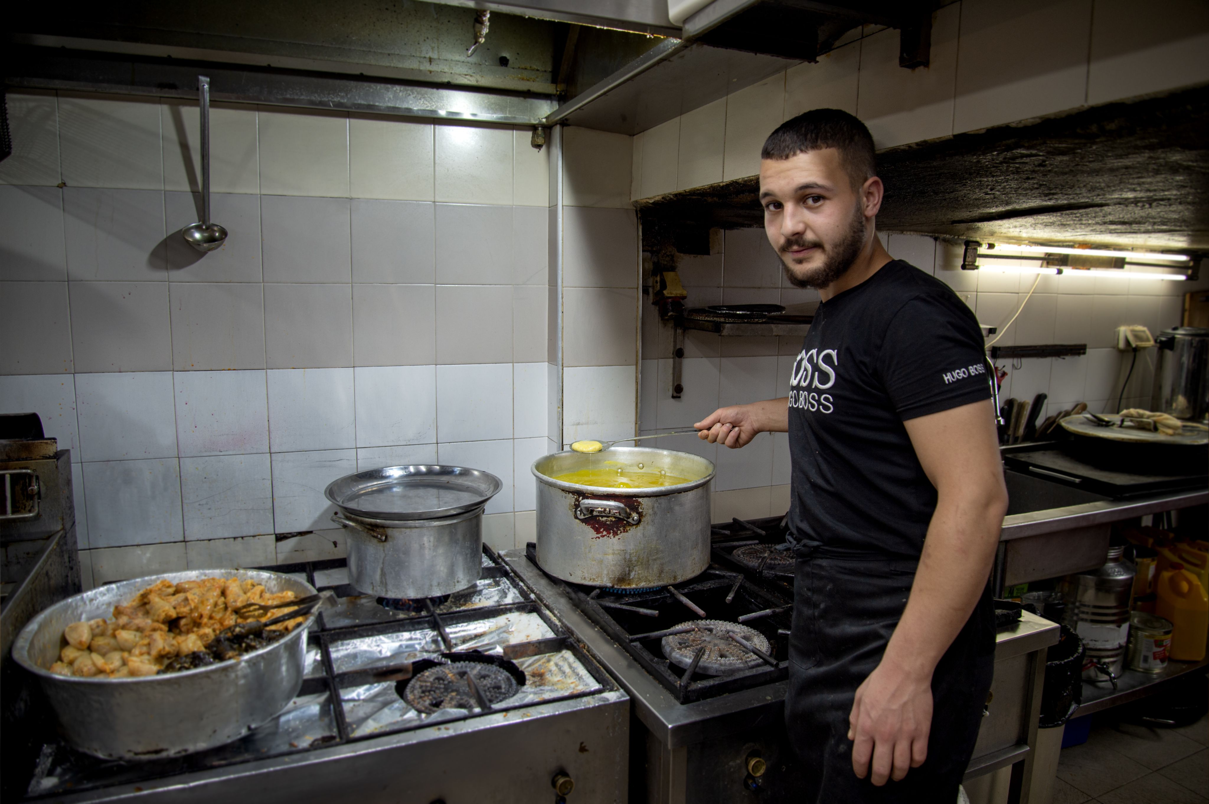 Colour photograph, indoors, man holding spoon looking towards camera, behind him are various kitchen utensils.