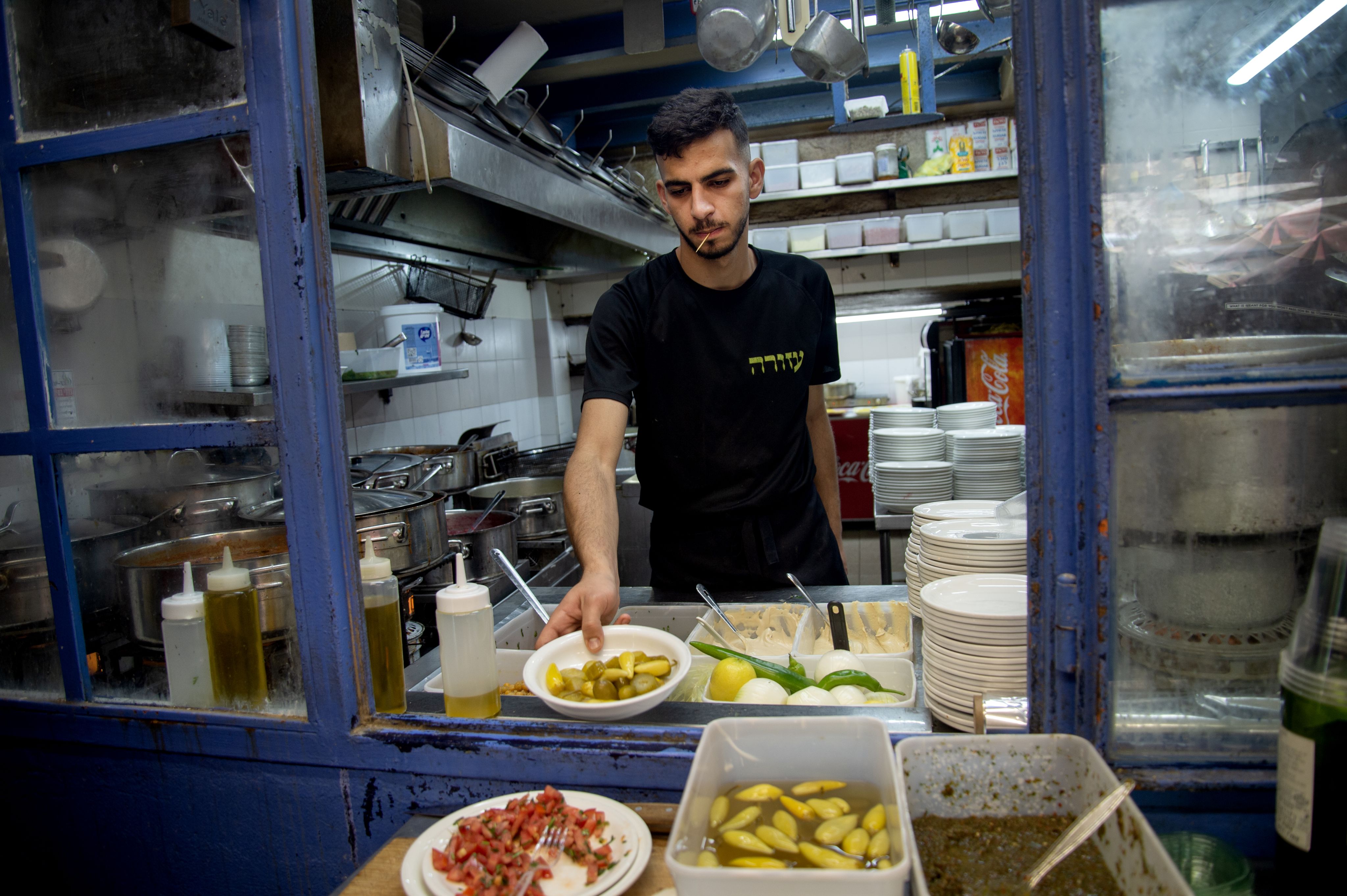 Colour photograph, indoors, man with toothpick handing food out of kitchen window, various spices in close up, various cutlery in kitchen in the background.