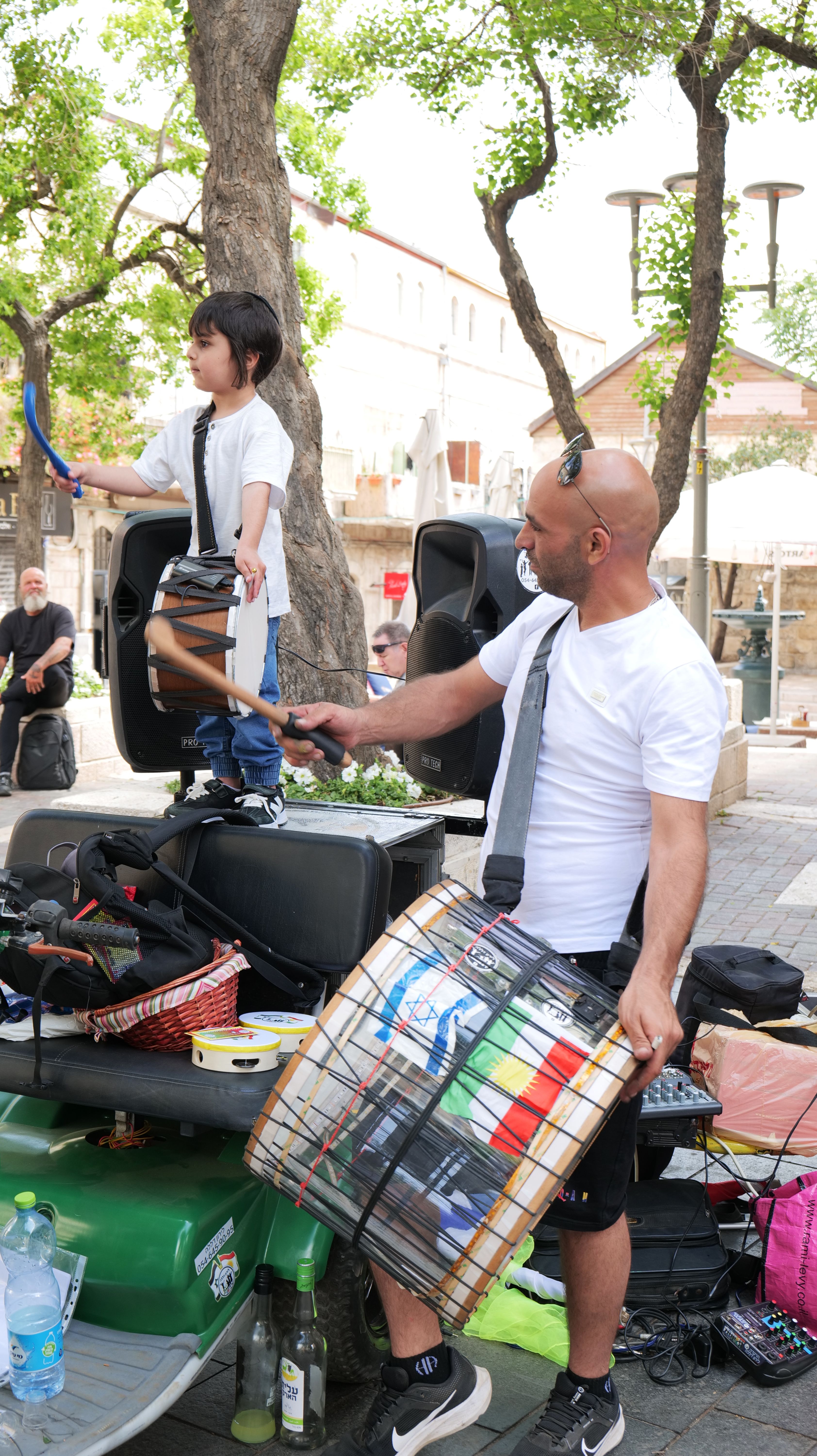 Colour photograph, outdoors, of men playing a large drum in the street and a child standing on cases playing a small drum.