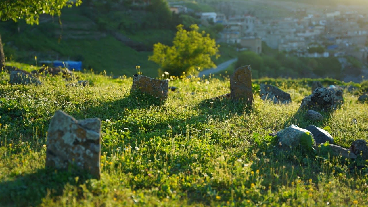 Colour photograph showing sunlight streaming into a field with small, crumbling gravestones. The landscape of a city is visible in the background.