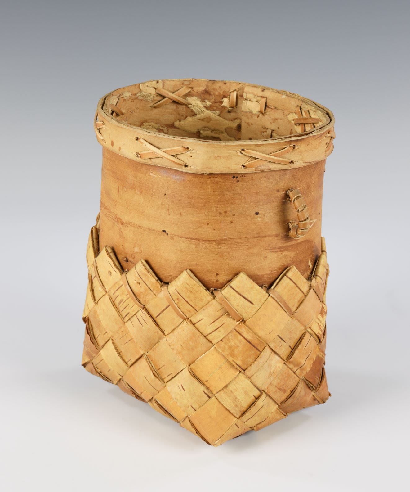 Birch bark container made of two different parts joined together. The top of the container has two small rounded handles and a protruding rim decorated with 'x' shape stitches.