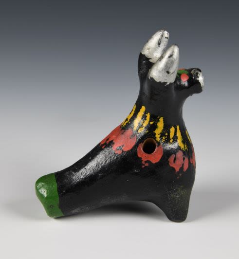 Whistle cow toy made of clay. The cow has a small head with nicely modulated horns and muzzle. The toy is painted in black enamel dyes with green forehead, red round eyes and silver horns with muzzle. The trunk is decorated with coffee bean like pattern in yellow and red.
