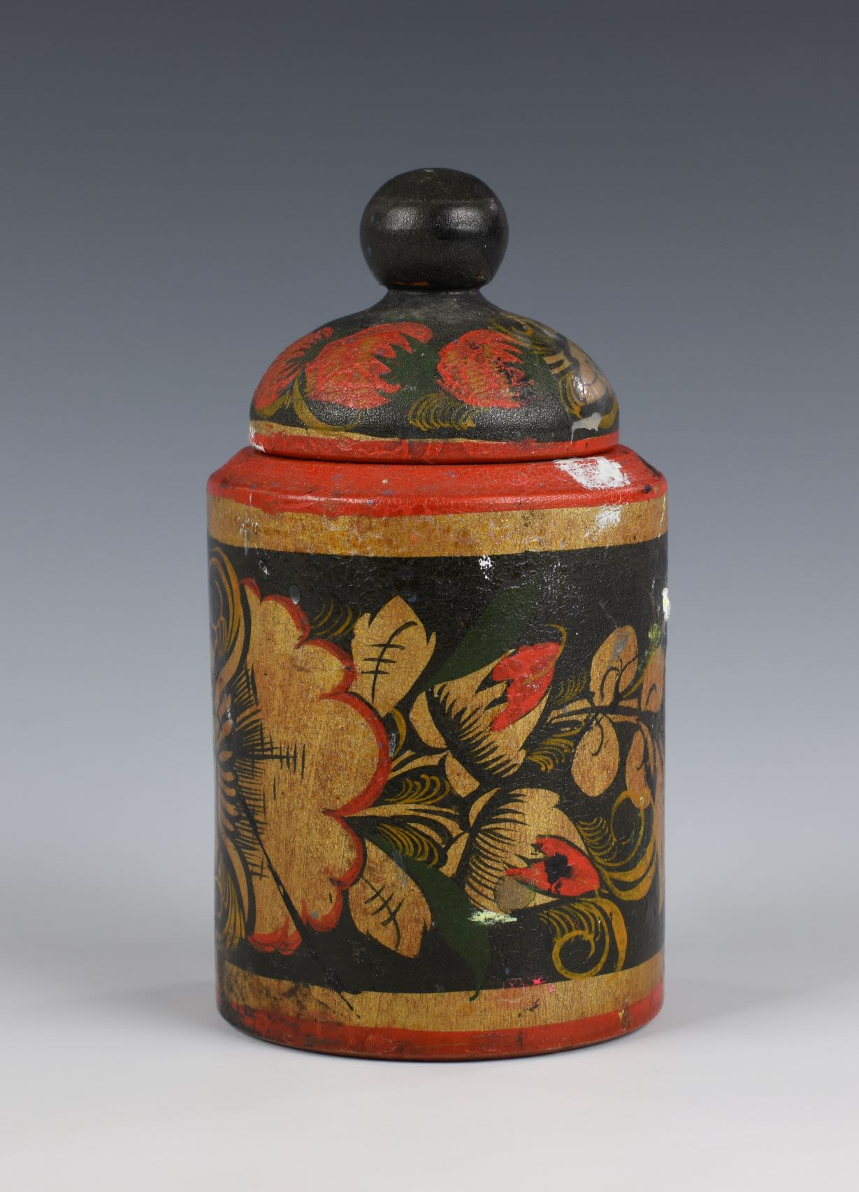 Lacquer wooden jar with the lid. The tube shaped jar with thin stripes of red and gold at the top and bottom. Wide central part decorated with floral patterns depicting a cluster of red rounded berries, gold flowers, gold leaves with elements of green grass on a black background. Inside of the jar is silver.