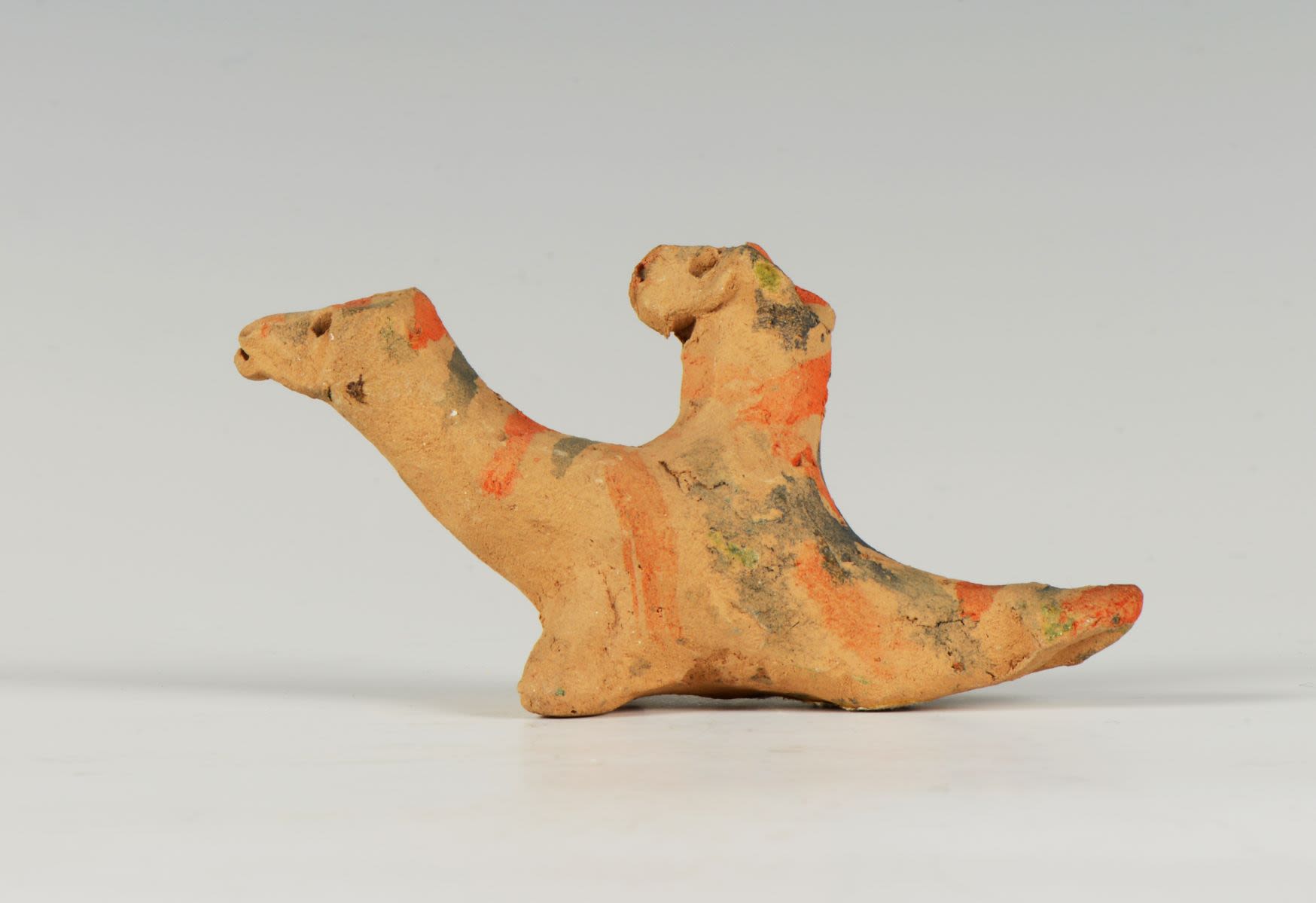 Clay toy of a mythical creature with two heads moulded in a very primitive manner. Decorated with a geometrical pattern of small holes and short horizontal stripes in blue and orange.