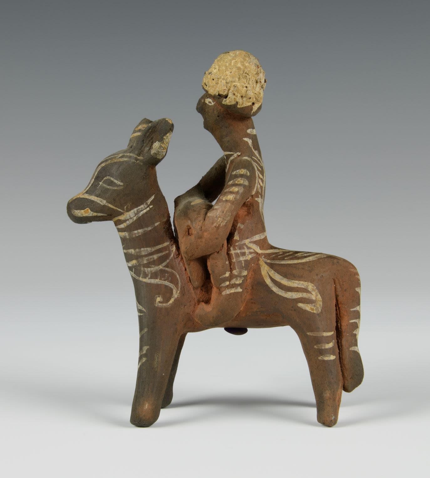 Clay toy of a figure riding a donkey and holding an animal in his lap. The toy is decorated with white geometric patterns.