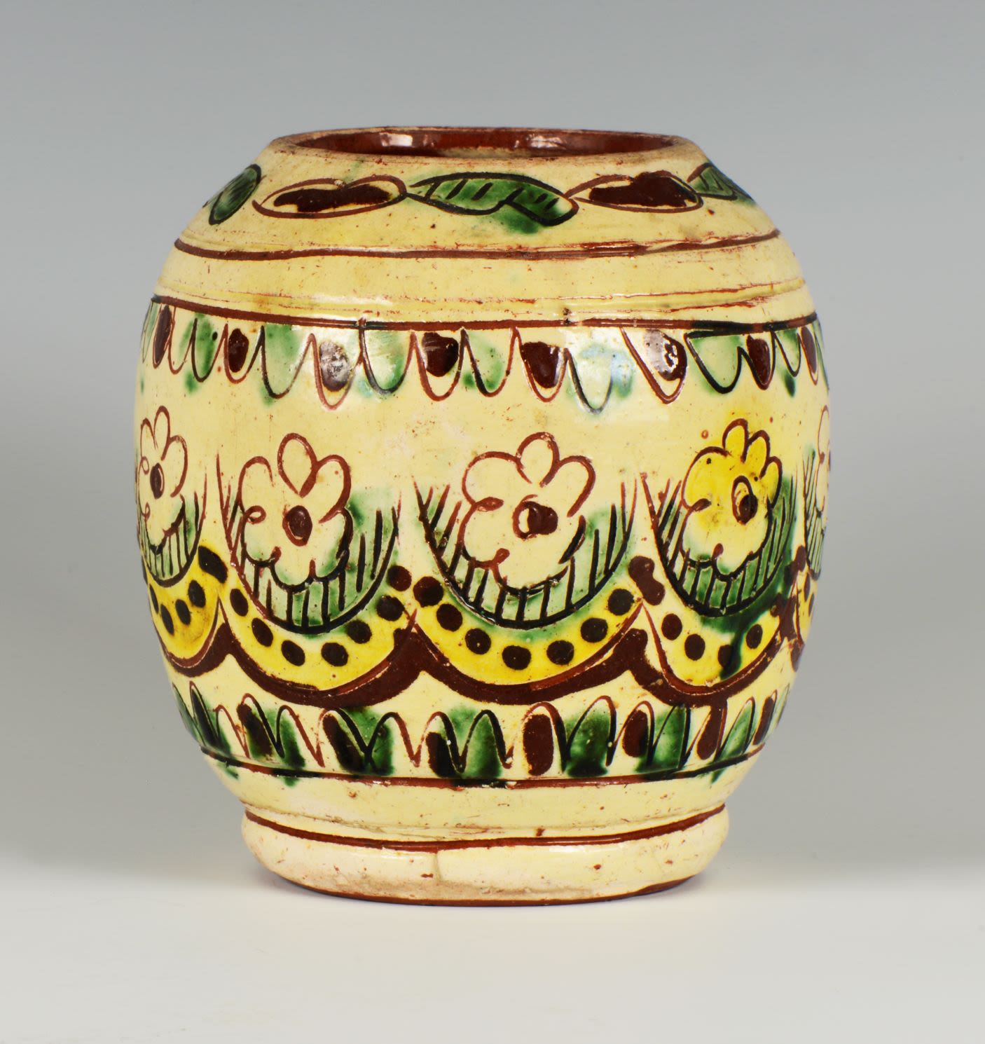 Ceramic jar decorated with narrow brown stripes and wavy pattern in green and brown,including flowers, leaves and stripes painted under the glaze in green, yellow and brown on a faded yellow background.
