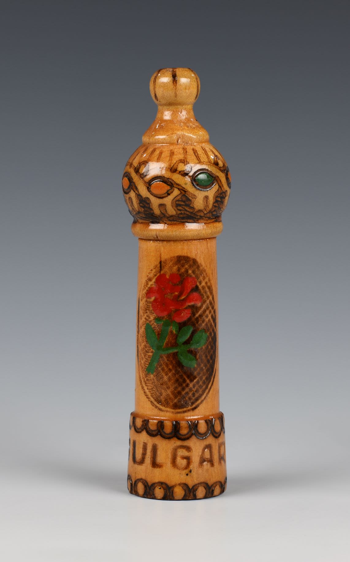 Wooden cylindrical case with a cover in the shape of a bottle fluted at the base with inscription 'BULGARIA' and rim with semi-circular pattern. The body decorated with coloured pyrography slightly tapers towards the base. The decoration depicts red rose with green leaves in an oval background and geometric ornament in brown, green and yellow.